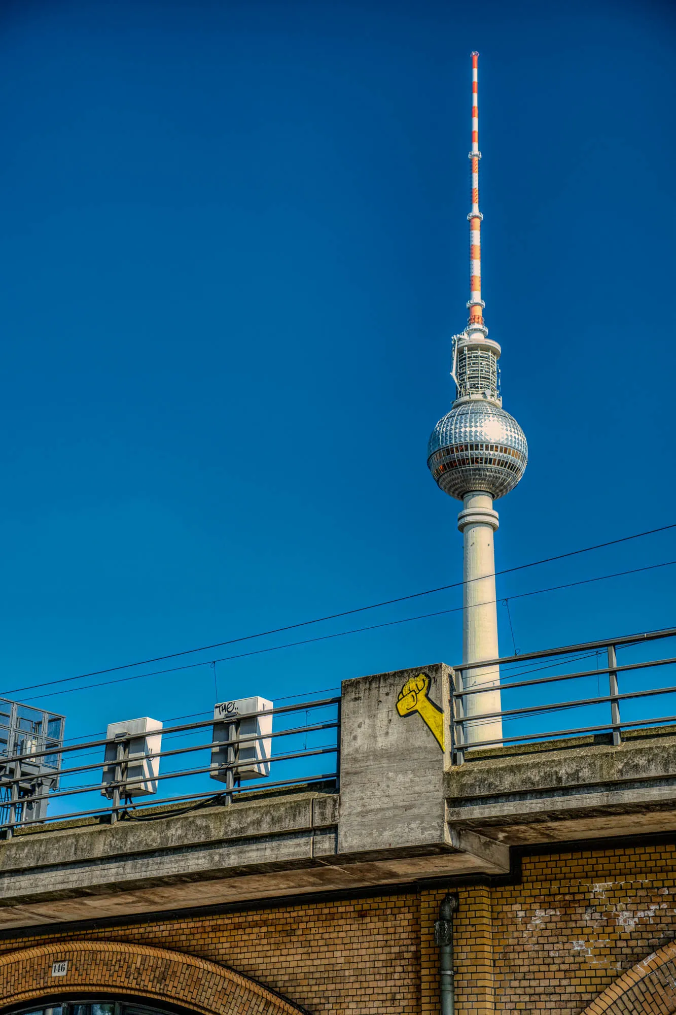 Fernsehturm Berlin behind the elevated subway tracks coming up behind a support with grafiti of a fist. Taken during golden hour with a clear blue sky