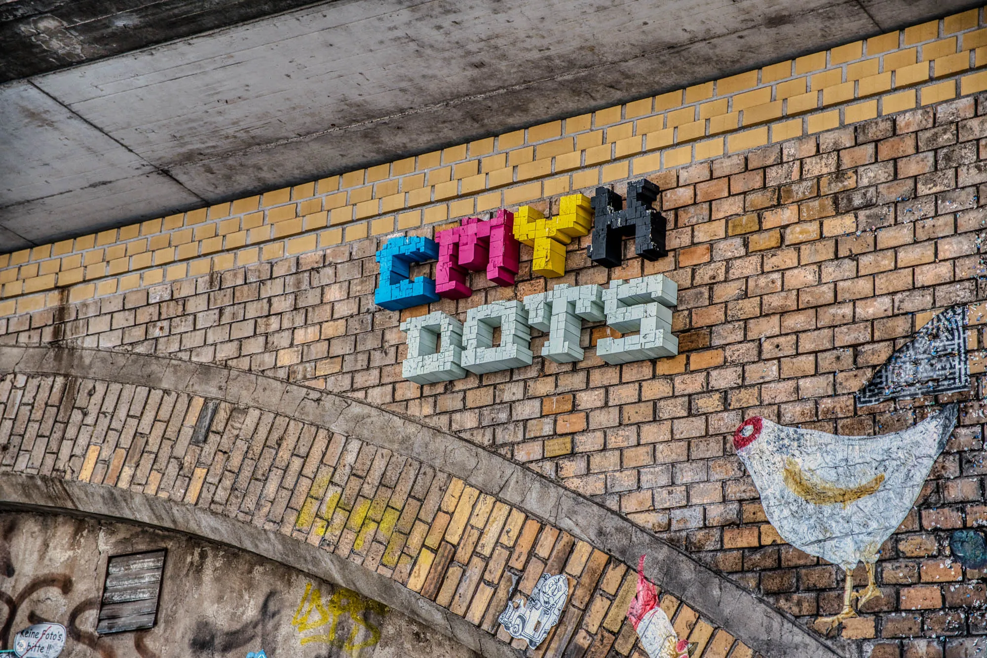 Close up shot of a brick wall under a bridge. There is graffiti of a chicken pointing to 3D graffiti that spells out "CMYK DOTS" with the C in cyan, M in magenta, Y in yellow, K in black, and DOTS in a cool white.