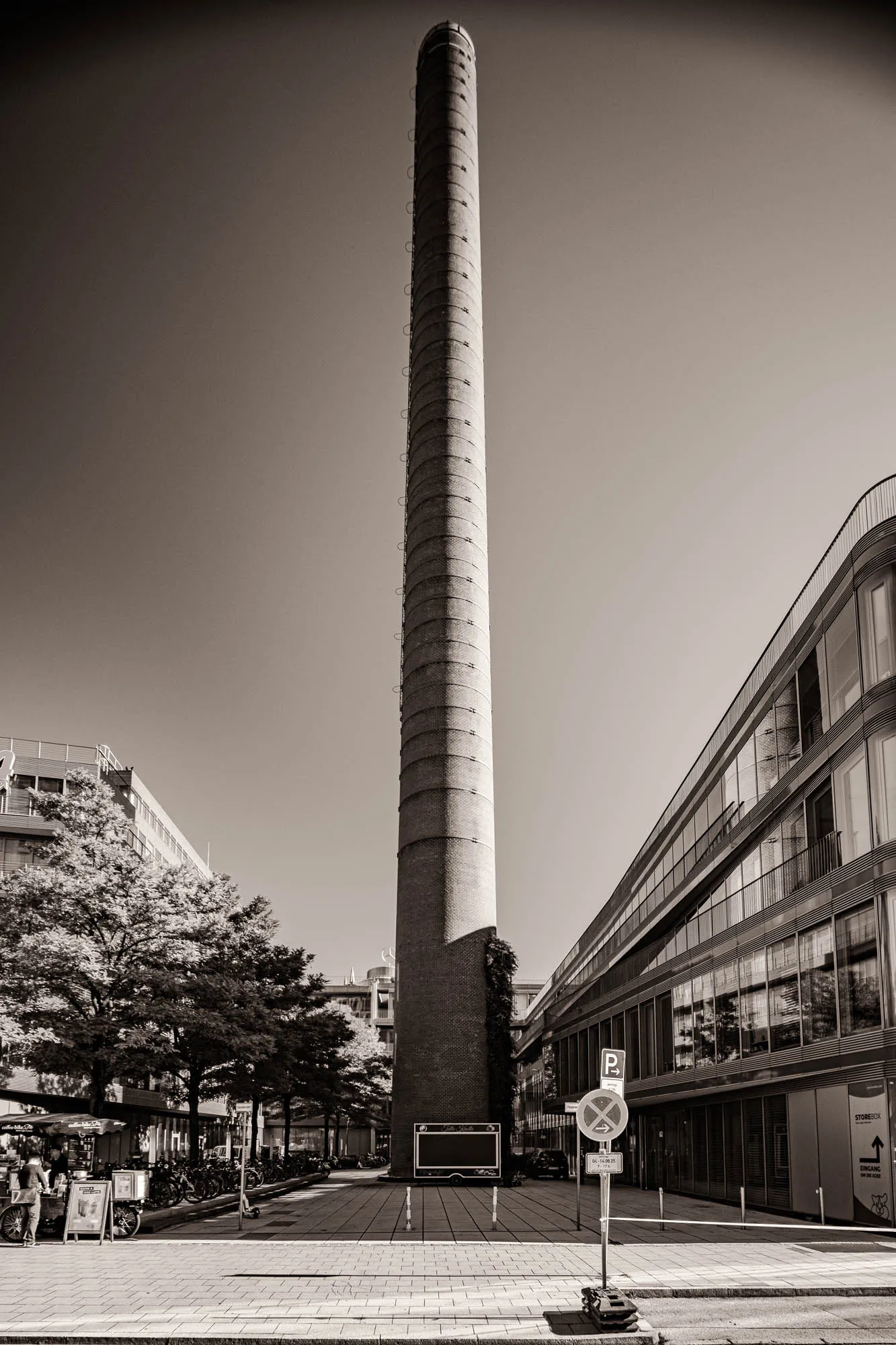 A black and white photograph presents a very tall, slender brick smokestack as the central element. The stack rises vertically from ground level, dwarfing the surrounding buildings. The smokestack is cylindrical with a ring of metal ladders running up its length, spaced at regular intervals. The surrounding cityscape includes modern buildings with glass facades that reflect the sky. To the left, leafy trees partially obscure a building, creating a sense of depth. The ground is paved and shows pedestrian areas, complete with parking signs, suggesting an urban environment. The sky is clear and bright. The monochrome palette accentuates the textures of the brick and the reflective surfaces of the buildings, enhancing the contrast between the old industrial structure and the modern architecture around it.