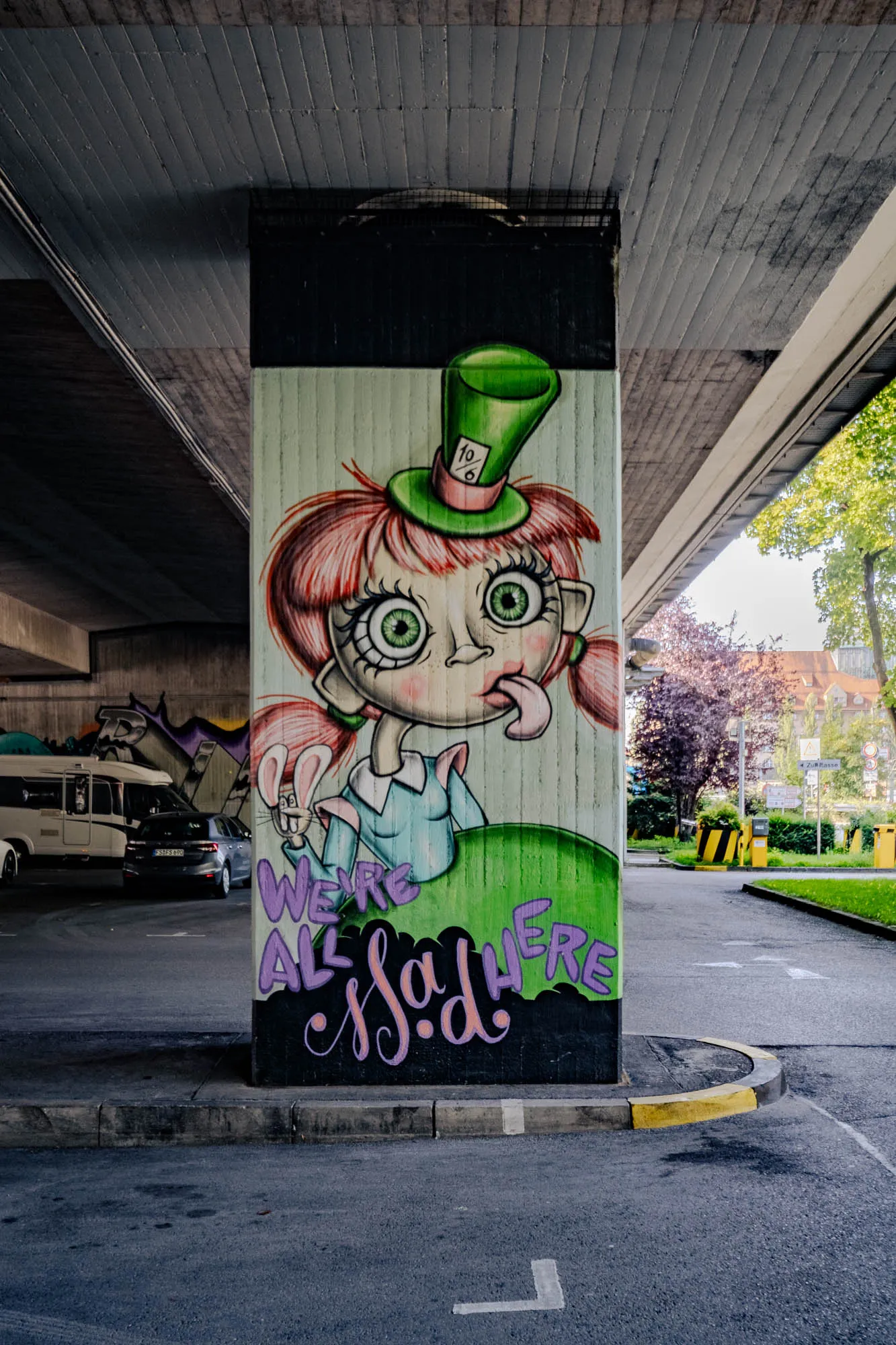 A painted concrete pillar supports an overpass, displaying a cartoonish image reminiscent of Alice in Wonderland's Mad Hatter, but in a quirky, original style. The character, appearing female, has pale skin, large, wide green eyes with thick black lashes, and playfully sticks her tongue out to the side. Her reddish-pink hair is styled in pigtails with a blunt fringe. She wears a tall, green top hat decorated with a '10/6' card on a pink ribbon. Below her, stylized lettering in purple and pink reads 'We're all mad here.' The pillar is painted light green above the writing and dark green below. The gray concrete overpass looms above, while a street scene with vehicles and greenery stretches beyond the pillar, indicating an urban or suburban environment.
