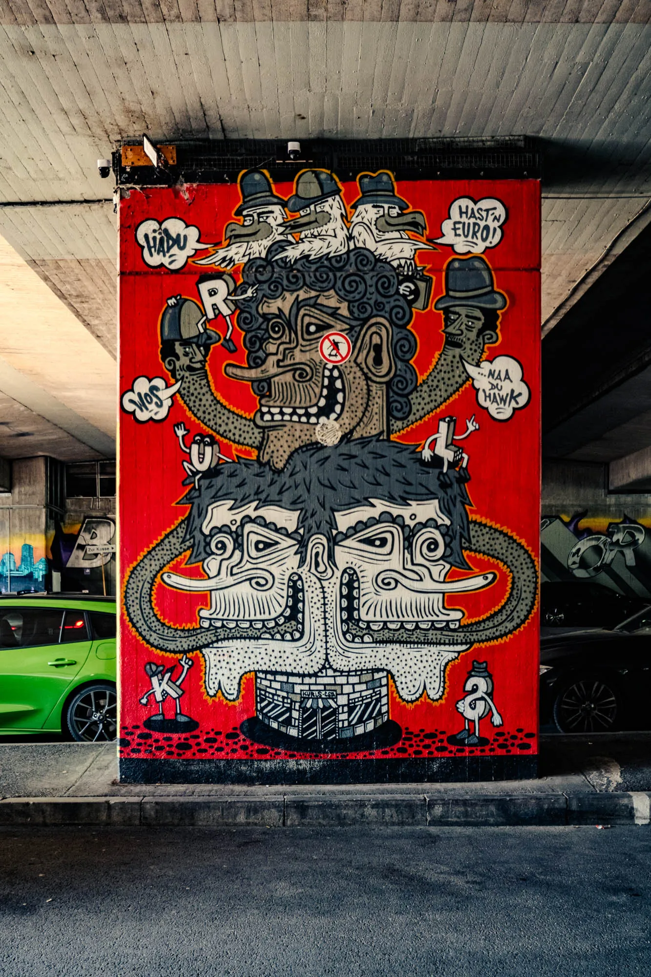 A large, vibrant graffiti mural dominates the frame, painted on a support pillar beneath an overpass. The pillar, primarily painted in a bold red, showcases a complex composition of cartoonish, anthropomorphic figures drawn in black and white with grey shading. At the top, three birds with fedoras perch, while below them, a central character with curly hair, a single visible eye with a prohibition sign over it, and an exaggerated, toothy grin commands attention. Two spindly arms extend from this character, each topped with a smaller, simpler figure wearing a hat. Beneath the central character, two stylized faces are conjoined, forming a mask-like shape with elongated noses and geometric patterns. The base of the mural features a brick-like pattern with the inscription 'Kyrlos.com'. Small, whimsical figures with stick-like limbs are scattered around the mural. The graffiti is partially obscured by the overpass structure above. To the left of the pillar, a bright green car is visible, while a dark-colored car is parked to the right. The ground in front of the mural is asphalt. The overpass's concrete construction is visible in the background, adding depth and context to the urban scene.