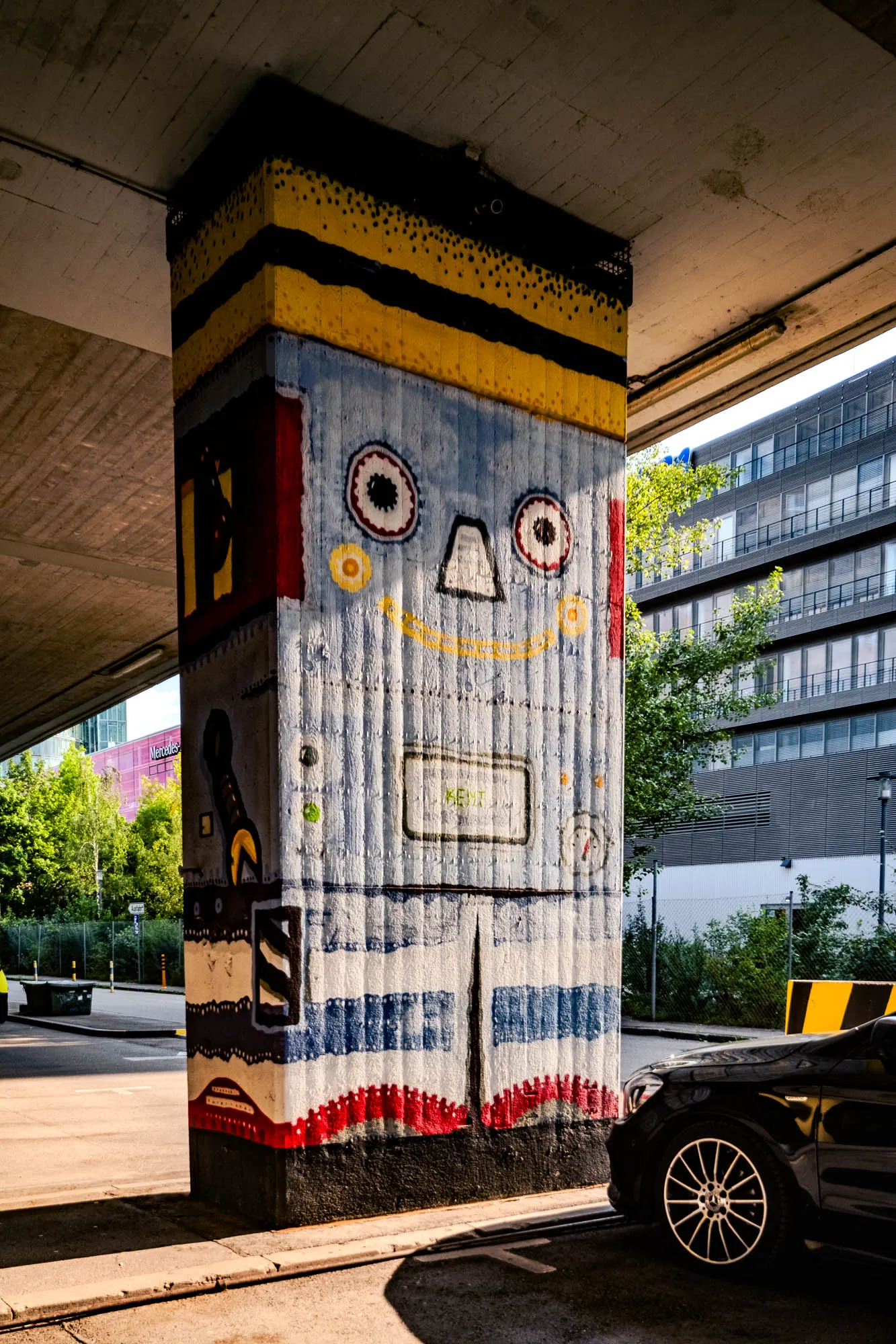 The photo captures a large, rectangular concrete pillar, prominently painted with a whimsical robot face, supporting an overpass. The top of the pillar is decorated with horizontal bands of yellow paint speckled with black dots, bordered by a thick, solid black stripe. The robot's face is painted in pale blue and white, featuring two large, round eyes with circular patterns, a simple triangle nose, and a rectangular screen-like mouth containing the word 'KENT'. A thin, curved yellow line suggests a smile. The lower section of the pillar has horizontal stripes of red, white, and blue. Along the left side of the pillar, a red stripe runs vertically, interspersed with abstract, dark painted shapes. To the right of the pillar, sunlight illuminates a street scene with modern buildings, lush green trees, and the front of a sleek, black car partially visible at the bottom of the frame.