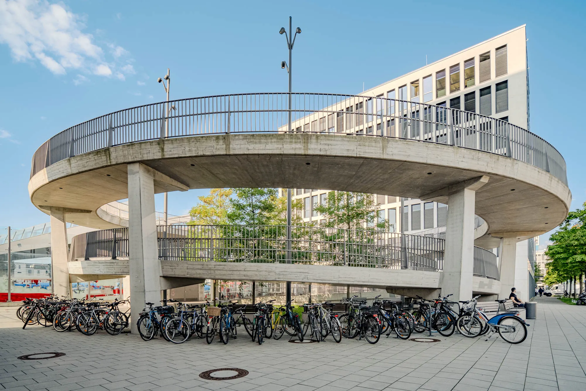 The image showcases a modern architectural scene under a partly cloudy sky. The focal point is a concrete, circular pedestrian walkway elevated above ground, supported by sturdy concrete pillars. The walkway is bordered by a metal railing that provides a visual break against the concrete. Underneath this circular structure, many parked bicycles of different colors, shapes, and sizes create a busy, urban feel. The ground is covered in a geometric pattern of grey tiles. In the background, a modern office building with many windows rises sharply with light walls, contrasting against the sky. The building's design features clean lines and a distinctive triangular shape. Between the walkway and the building, a line of trees with lush green leaves adds a touch of nature. Several thin lampposts are visible, reaching up towards the sky along the side of the bridge.