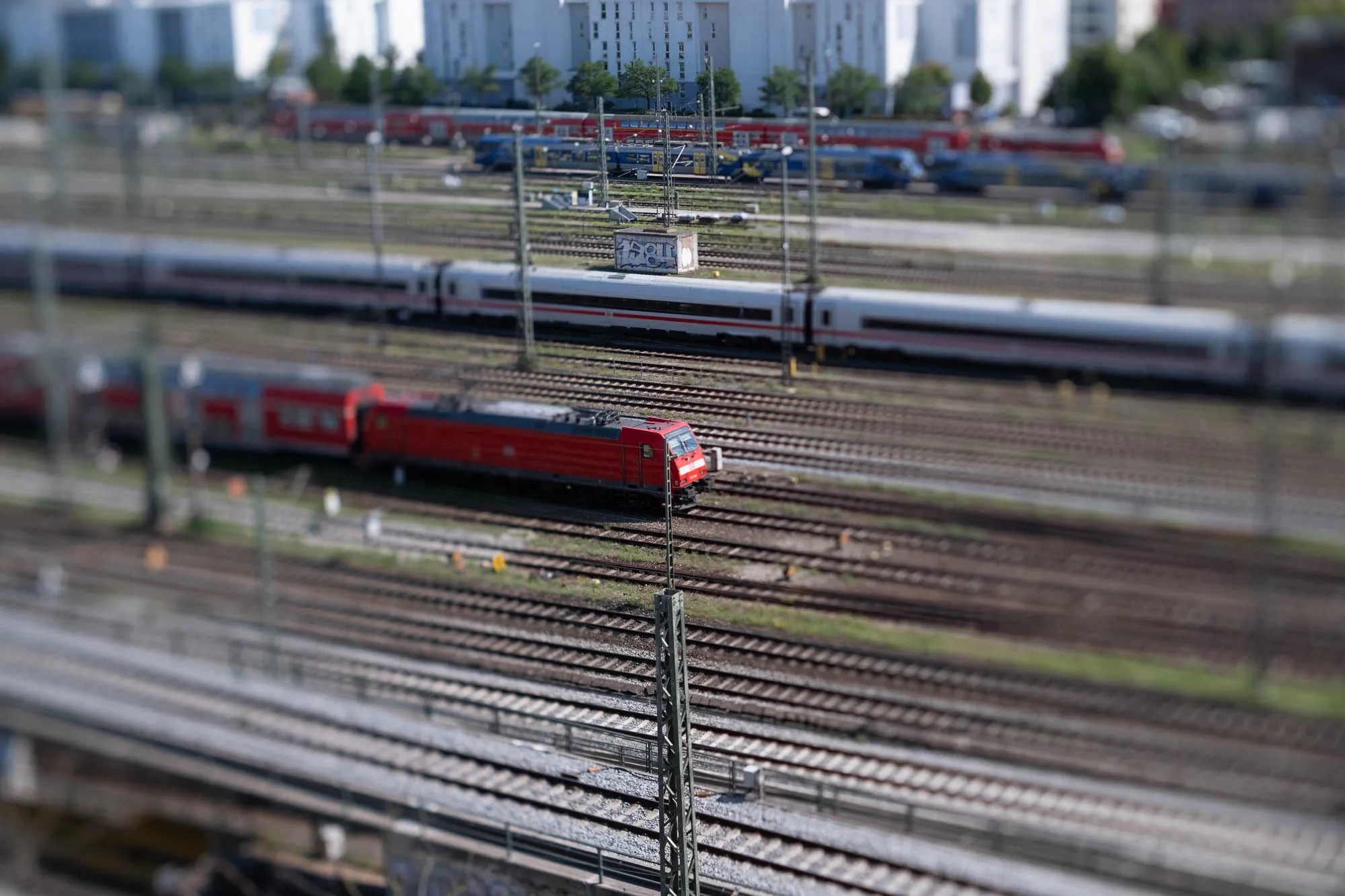The image presents a wide-angle view of a train yard, meticulously composed with a tilt-shift effect, rendering elements out of focus to mimic a miniature scale. Multiple parallel train tracks dominate the foreground and middle ground, converging towards a distant backdrop of tall, modern buildings. A bright red locomotive stands out prominently, slightly blurred, traversing one of the tracks. Behind it, a long, silvery-white train stretches horizontally across the frame. In the far distance, more trains, some blue and some red, are parked on different tracks, adding depth and complexity to the scene. Overhead wires and metal poles punctuate the space above the tracks, creating a linear framework. The buildings in the background are tall and rectangular, their windows reflecting the sunlight. The overall color palette is muted with dominant hues of gray, brown, red, and white, creating a sense of vastness and intricate infrastructure. The tilt-shift effect adds a sense of whimsy and artistry to the image, emphasizing the miniature-like quality of the scene.