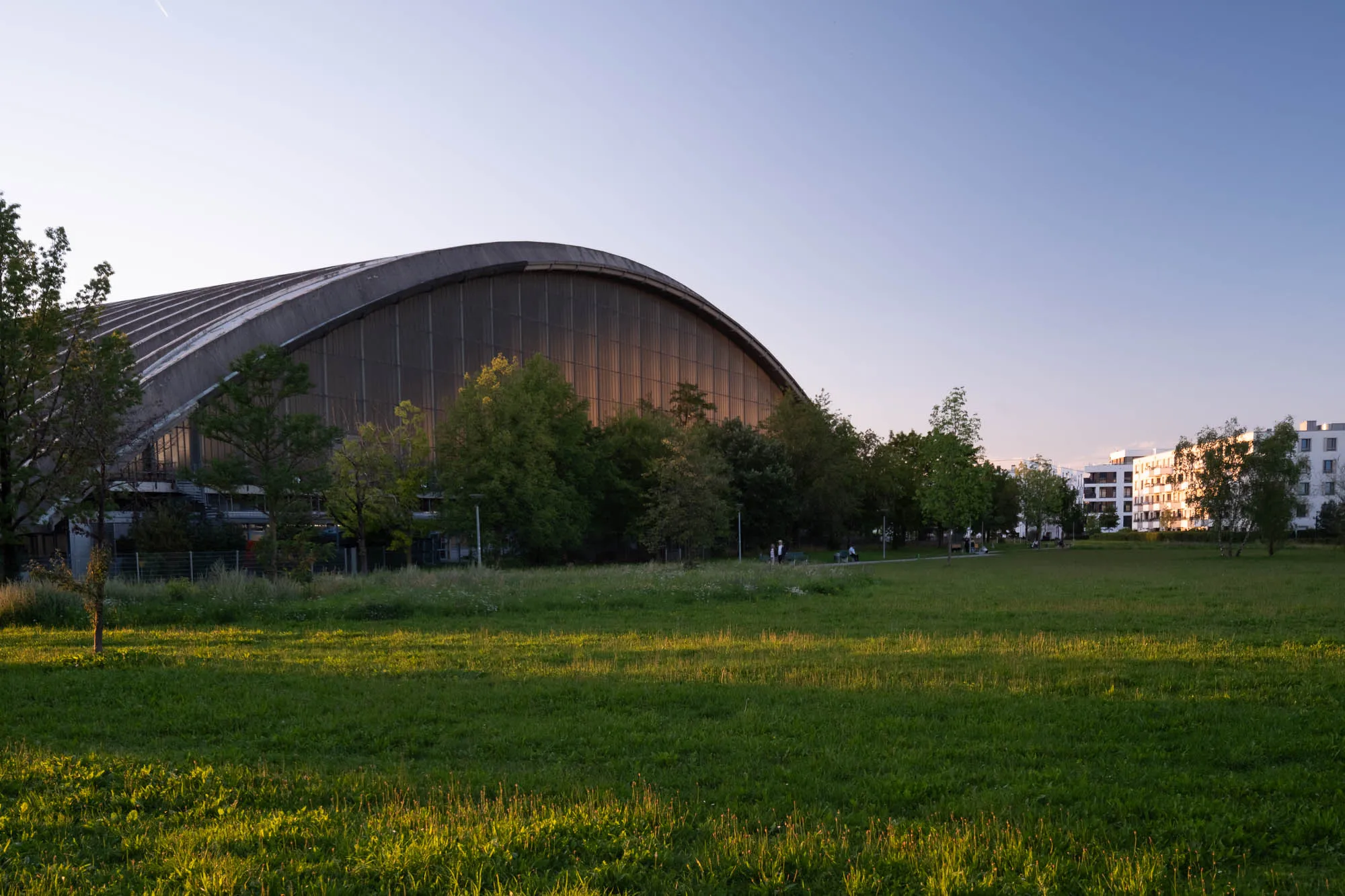 The image shows a large, arched building situated behind a lush, green field. The building has a distinctive, rounded roof that sweeps gracefully across the horizon. It appears to be constructed from metal paneling, giving it a textured, industrial feel. Sunlight reflects off the building's surface, highlighting the panels and creating a warm glow. Several mature trees partially obscure the building, adding a touch of nature to the scene. The field in the foreground is a vibrant green, with grass blades standing tall. A path winds through the field, leading towards the building and inviting exploration. In the background, several white apartment buildings can be seen, suggesting an urban or suburban setting. The sky is a clear blue, gradually fading to a pale lavender near the horizon. The overall impression is one of tranquility and quiet beauty, with the building standing as a landmark within a natural setting.
