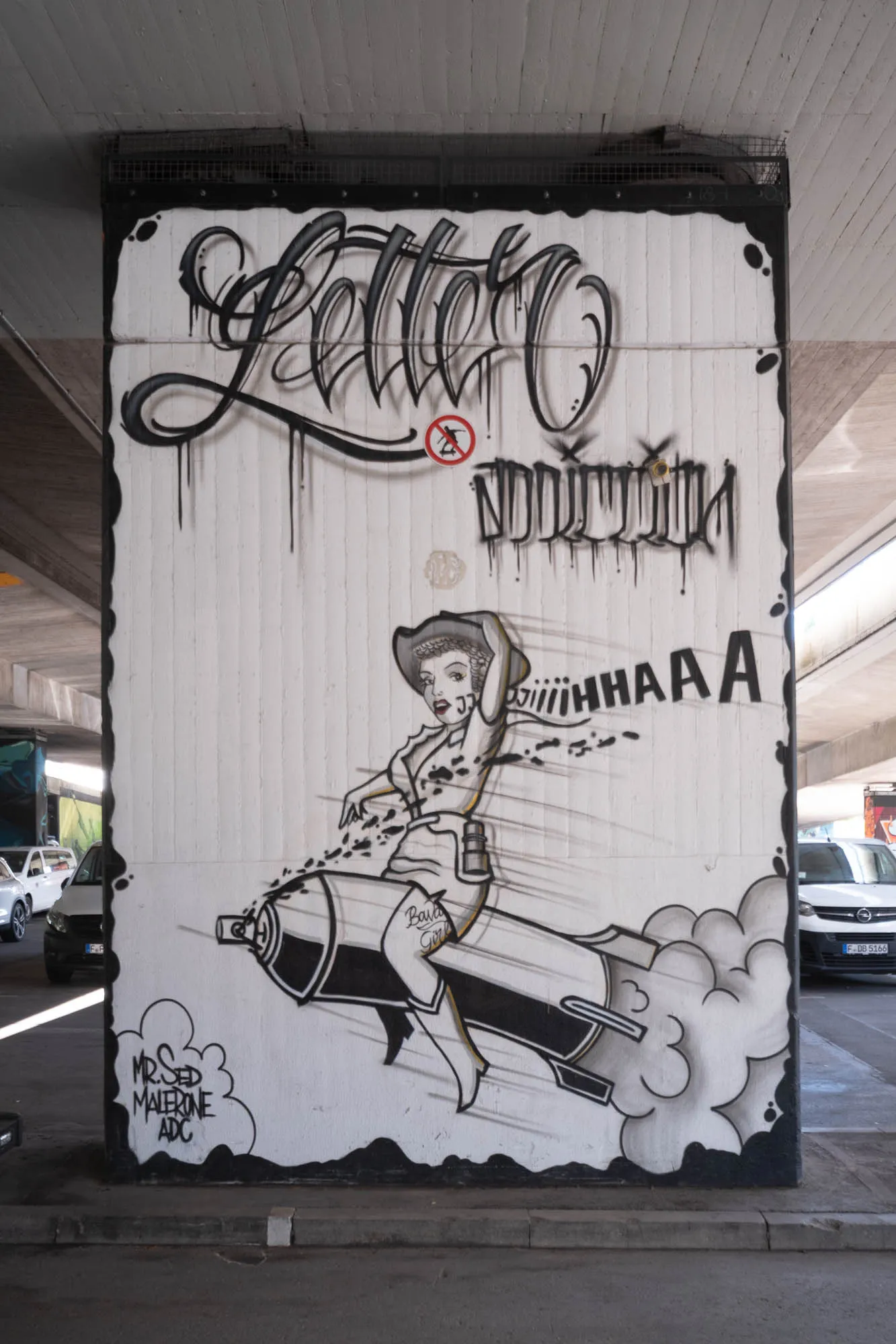 A large graffiti mural, predominantly in black and white, covers a concrete pillar under what appears to be a bridge or overpass. The artwork features a caricature of a woman, seemingly inspired by a retro pin-up style. She sports a cowboy hat and is depicted riding a large, cartoonish bomb that resembles a lipstick tube with fins. Smoke billows out from the back of the bomb, suggesting it's in flight. The woman raises one arm in the air with an expression of exhilaration or perhaps a slightly crazed grin, with stylized lines indicating speed and movement around her. Text is incorporated into the mural; above the woman, 'letter' is written in a fluid, calligraphic style, and another indecipherable word is underneath. Large, drawn letters spell out 'iiiiiHHHAAAAA' following the bomb.  The mural is outlined with a black border, and additional text at the bottom designates the artist, including 'MR.SED MALERONE ADC'. In the background, partially visible behind the pillar, are cars and other elements suggesting an urban environment.