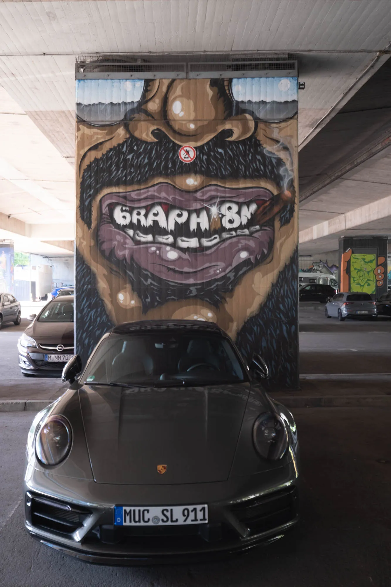 A gray Porsche 911 is parked in an urban parking garage, positioned directly in front of a large, whimsical mural painted on a concrete support pillar. The mural depicts a cartoonish face with a thick, dark beard, exaggerated lips, and a cigar clenched between its teeth. The word "GRAPHISM" is embedded within the teeth. A small, circular sign with a crossed-out figure presumably indicating no smoking is also present on the mural. The car's license plate, "MUC SL 911," is clearly visible. Other vehicles are parked in the background, and overhead concrete structures create the typical parking garage setting.