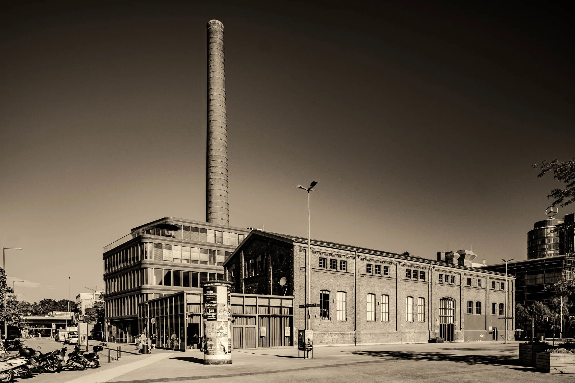 The sepia-toned photograph presents a cityscape where modern and historical architecture converge under a clear sky. In the center of the frame, a long, rectangular brick building dominates the view, its facade punctuated by regularly spaced arched windows. To the left, a more modern structure rises, featuring a grid of glass and metal, suggesting an office or residential building with multiple stories. A massive, cylindrical smokestack stands tall behind these structures, rising high into the sky. In the foreground, a paved area extends across the bottom of the image, adding a sense of depth and scale. A number of motorcycles are parked to the left side of the frame, near what seems to be a sidewalk, adding a sense of urban activity. Streetlights stand as vertical elements in the scene, while the presence of trees and the Mercedes-Benz logo on top of a modern building on the right edge suggest a contemporary urban environment. The lighting is bright and even, highlighting the textures of the brick and glass.