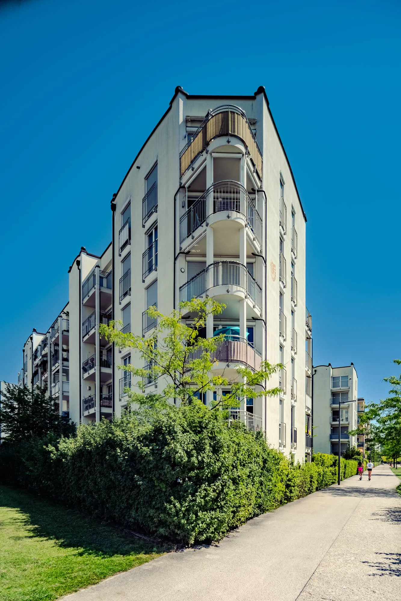 The image shows a multi-story, modern apartment building on a sunny day. The building has a light, creamy white facade, and features rows of balconies with curved metal railings. Windows are regularly spaced along the building's face, some with closed blinds. A vibrant green hedge borders the building at ground level, adding a natural element to the urban setting. Beyond the building and to the right, a paved walkway extends into the distance, lined with trees providing shade. Two people can be seen walking on the path, suggesting a peaceful and active environment. The sky above is a clear, bright blue, providing a stark contrast to the lighter tones of the building.