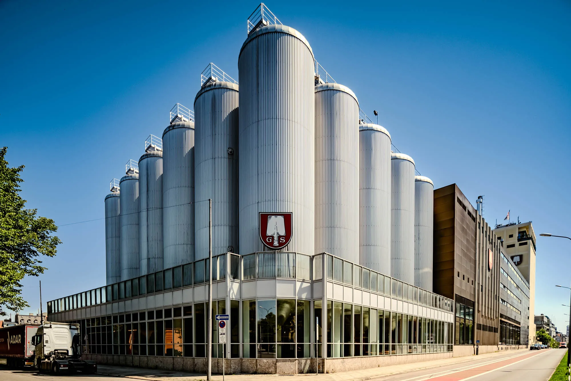 The image shows the distinctive Spaten-Franziskaner-Bräu in Munich. The building is dominated by a cluster of about ten towering, cylindrical, stainless steel silos. Each silo has a slightly domed top, and is vertically corrugated. At the peak of each silo sits a small railed platform. These silos rise from behind a two-story building with large windows framed in white that wrap around the corner of the structure, giving a view of the inside. A sign with the brewery's logo is visible in the middle on top of the building; it features a white and red shield with the head of a white shovel head on it, with G and S on either sides of where the head would meet the handle. To the right of the silos, the building transitions to a darker brown, more rectangular structure. The sky above is a clear, bright blue, indicating a sunny day. A few trees with lush green leaves are visible to the left, and a truck is partially visible near the street corner. The street stretches in front of the building, with a red lane marked on the side, indicating a bike path.