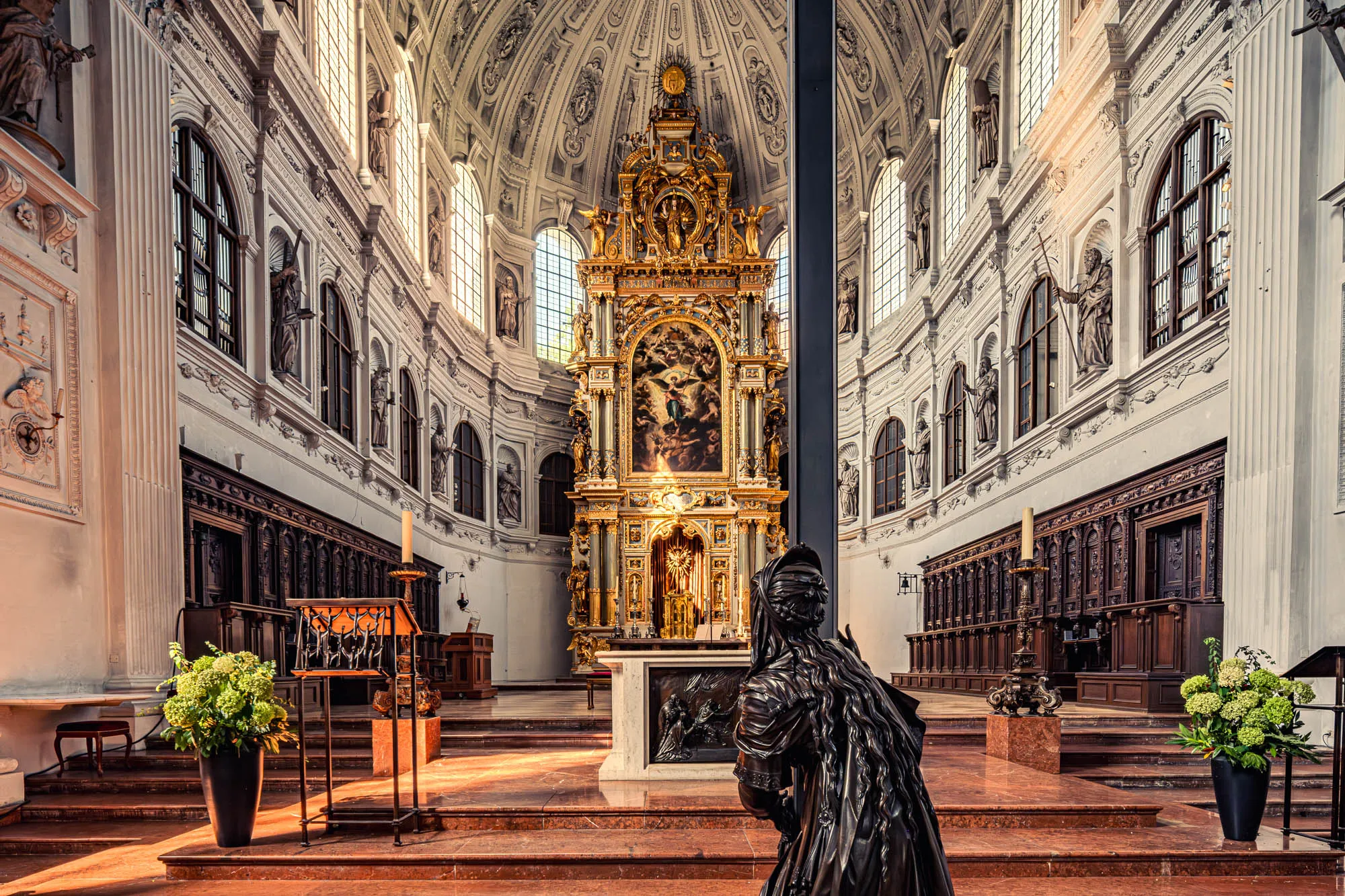 The image shows the interior of a grand, ornate church. The dominant colors are warm reds and browns contrasted with cool whites and creams. In the foreground, a dark, metallic statue of a kneeling figure faces away from the viewer, drawing the eye into the church's depths. This figure has what appears to be a cloth draped over its head and shoulders. The church floor is composed of light brown and light-red marble tiles, arranged in a stepped pattern leading towards the altar. On either side of the foreground, large black vases filled with light-green hydrangea-like flowers flank the steps. Dark wooden pews line both sides of the church's interior, adding to the sense of traditional architecture. Along the walls, numerous stone statues are positioned in niches beneath tall, arched windows. Natural light streams through these windows, illuminating the space. The focal point is the massive golden altar, its intricate carvings catching the light. Above the altar, the church's dome rises, adorned with detailed plasterwork and culminating in a central point above. The overall impression is one of great size, rich detail, and solemn beauty.