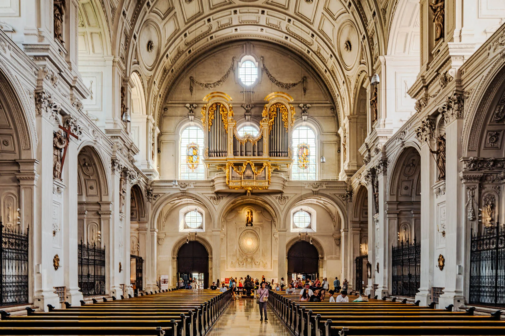 The image presents the interior of a grand, ornate cathedral. The perspective is from the rear, looking towards the altar. Rows of wooden pews stretch forward on either side of a gleaming marble aisle, disappearing into the distance. Sunlight streams through arched windows high above, illuminating the light beige walls and the richly decorated architecture. Tall, fluted columns flank each side, creating a series of arched openings that lead to side aisles. Iron gates separate the side aisles from the main sanctuary. Above the altar, a magnificent golden pipe organ sits beneath a central arched window, framed by intricate carvings and sculptures. Numerous people are visible scattered throughout the pews and along the aisle, some standing, others seated, creating a sense of activity within the vast space. The overall impression is one of grandeur, light, and meticulous detail, reflecting the historical and religious significance of the cathedral.