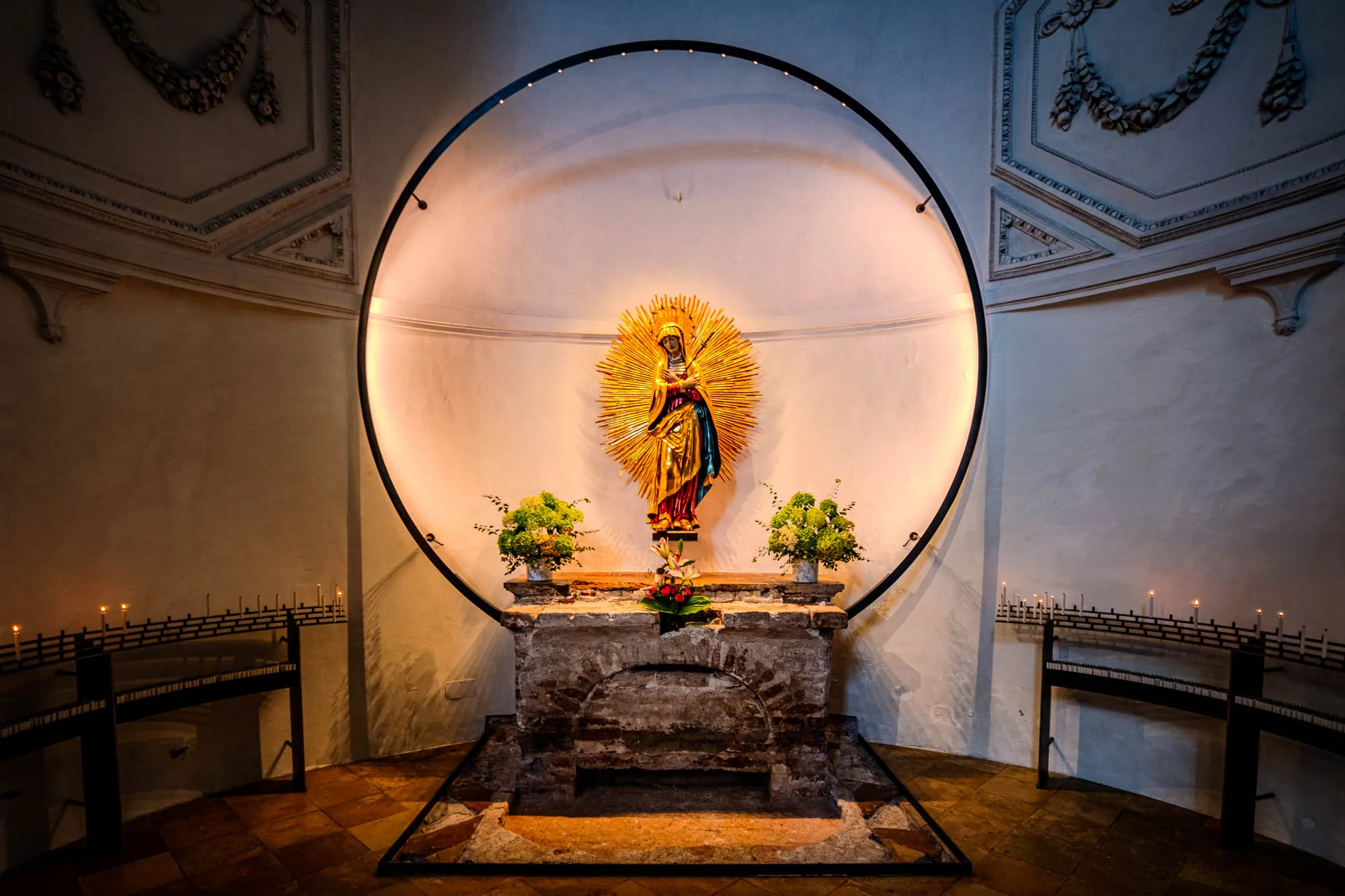 The image shows a niche in what appears to be a chapel or church, with a statue of the Virgin Mary as the focal point. The statue is positioned on a stone altar, illuminated by warm light that emanates from behind it, creating a halo effect. The statue itself appears gilded, with a golden, sunburst-like radiance extending outward, drawing the eye. The statue wears a blue shawl, and a long red dress, both with gold details. Flanking the altar on either side are small vases filled with greenery, providing a touch of natural color. Surrounding the altar is a large, black metal circle, which adds a modern geometric element to the otherwise classic and religious setting. On either side of the niche, are black wrought-iron candle racks each holding several lit candles. The walls of the chapel are a soft white, with intricate plaster detailing on the ceiling adding a touch of ornate texture. The floor is tiled with a mixture of dark and light stone, creating a patterned surface. The overall ambiance is one of reverence and peace, with the play of light and shadow adding depth and dimension to the scene.