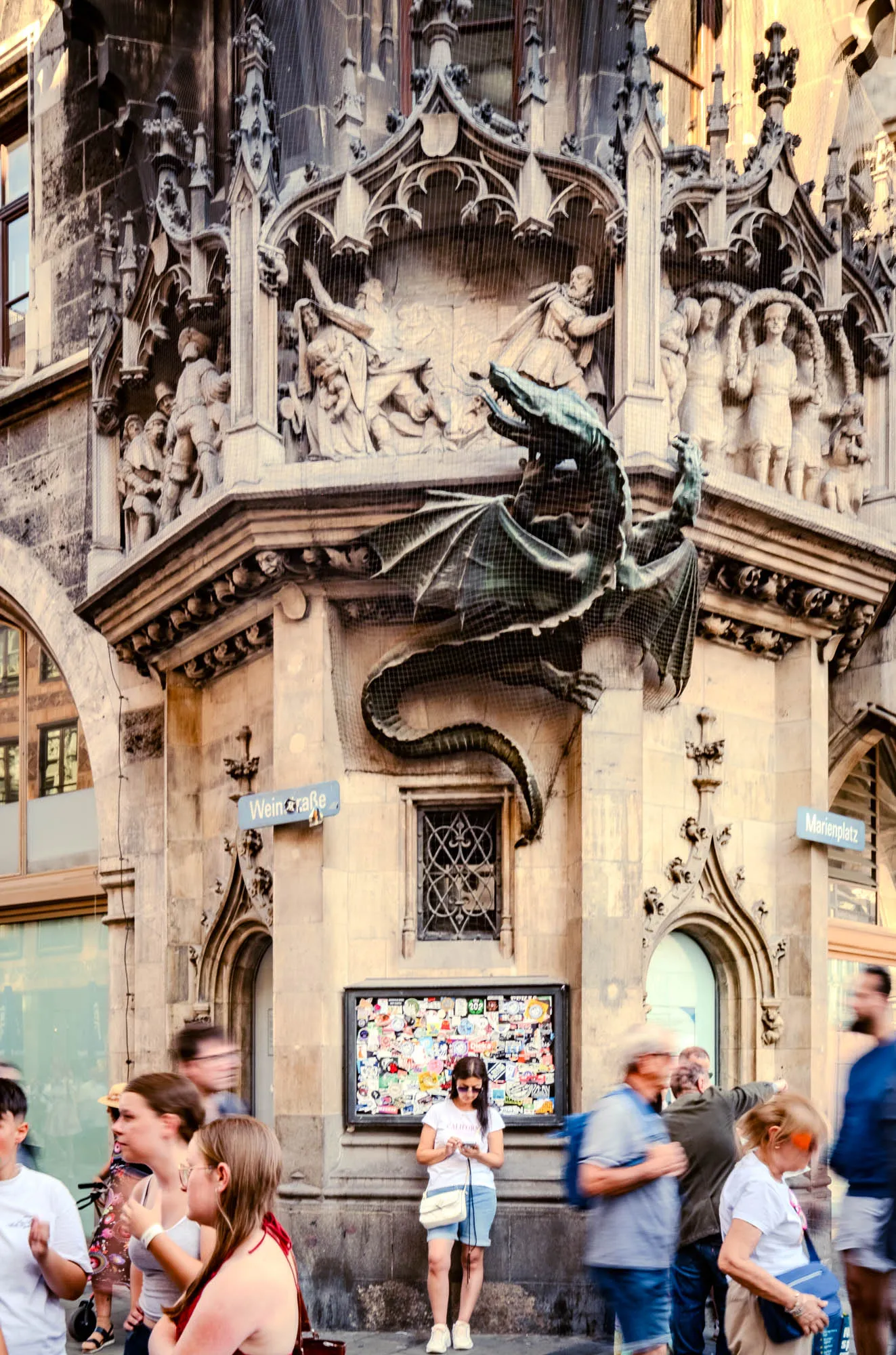 The image captures a bustling street scene in Munich, Germany, focusing on a detailed architectural corner of a building adorned with historical sculptures and a dragon statue. The corner is constructed of light-colored stone, elaborately carved with gothic arches, sculpted figures depicting historical scenes, and small windows. A large, three-dimensional dragon statue, made of a dark, possibly bronze-like material, is mounted prominently on the corner. The figures appear to be scenes of battle or historical events. Street signs for 'Weinstraße' and 'Marienplatz' are affixed to the building. Below the corner, on the street level, stands a young woman with long brown hair, wearing a white t-shirt, denim shorts, and sneakers. She has sunglasses perched on her head and is engrossed in her phone. Behind her is a bulletin board or display case covered in numerous stickers. Blurry figures of pedestrians pass by, giving a sense of motion and the lively atmosphere of the city.