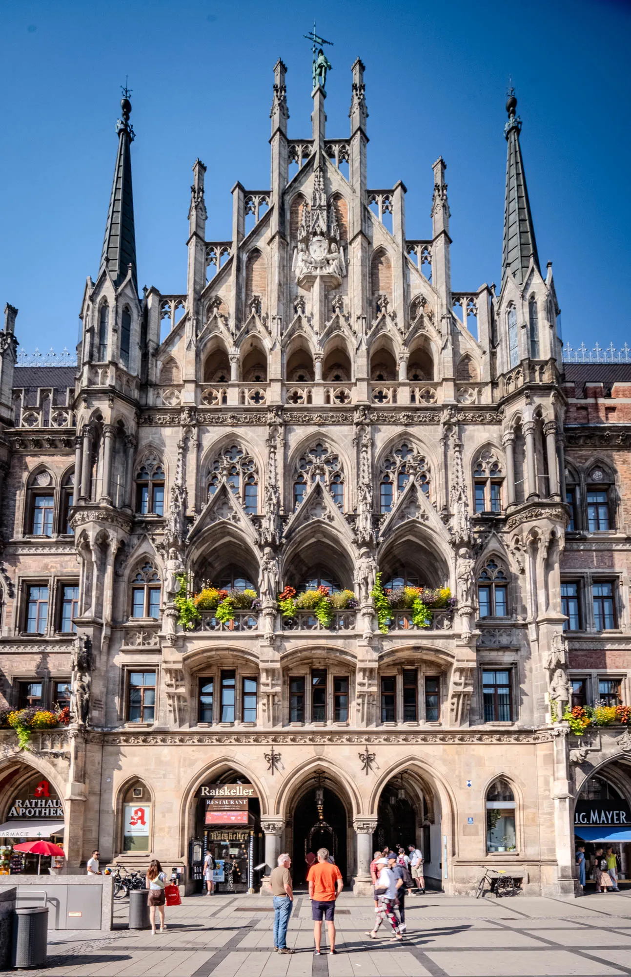 The image showcases the New Town Hall (Neues Rathaus) in Munich, Germany, under a bright, cloudless blue sky. The building is a grand example of Neo-Gothic architecture, constructed of light-colored stone. Its facade is intricately detailed with elaborate carvings, arches, and decorative elements. Several sharp spires rise from the building's roof, adding to its imposing presence. The building's façade has several stories, each one adorned with rows of windows and balconies. Bright flowers spill from the balconies on several levels. On the ground level, arched entryways lead into shops and restaurants, with people walking and standing nearby. A pharmacy and a restaurant with signage are also visible near the base of the building. The scene is brightly lit, casting shadows that emphasize the building's architectural details. A paved area in front of the building shows pedestrians walking about.