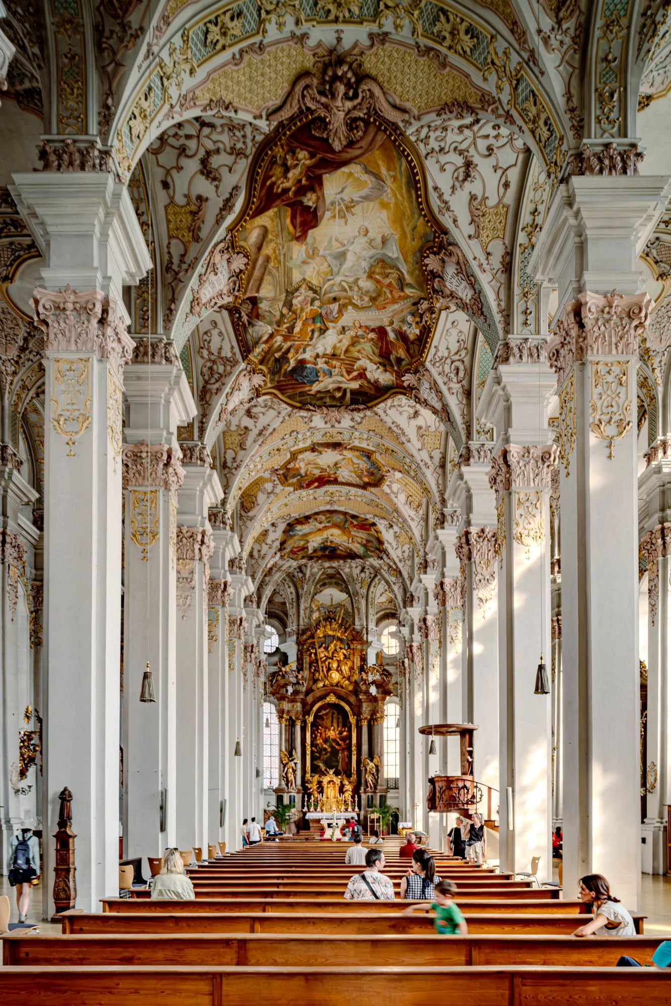 The image shows the breathtaking interior of a grand church, viewed from the back of the nave looking towards the altar. The perspective emphasizes the length and height of the space, creating a sense of awe and reverence. Rows of dark wooden pews, mostly occupied by individuals and small groups, stretch from the bottom of the frame towards the altar. Flanking the pews on either side are tall, imposing, white columns that reach to the elaborately decorated ceiling. These columns feature ornate gold and beige detailing on their capitals. The ceiling is a masterpiece of Baroque art, adorned with intricate paintings depicting religious scenes, framed by swirling patterns, gilded trim and decorative plasterwork. Sunlight streams in through windows high above, casting soft shadows and highlighting the rich details of the artwork. At the far end of the nave, the golden altar stands as the focal point, its details somewhat softened by the distance but still conveying a sense of grandeur and importance. The overall impression is one of magnificent architecture, artistry, and spiritual tranquility.