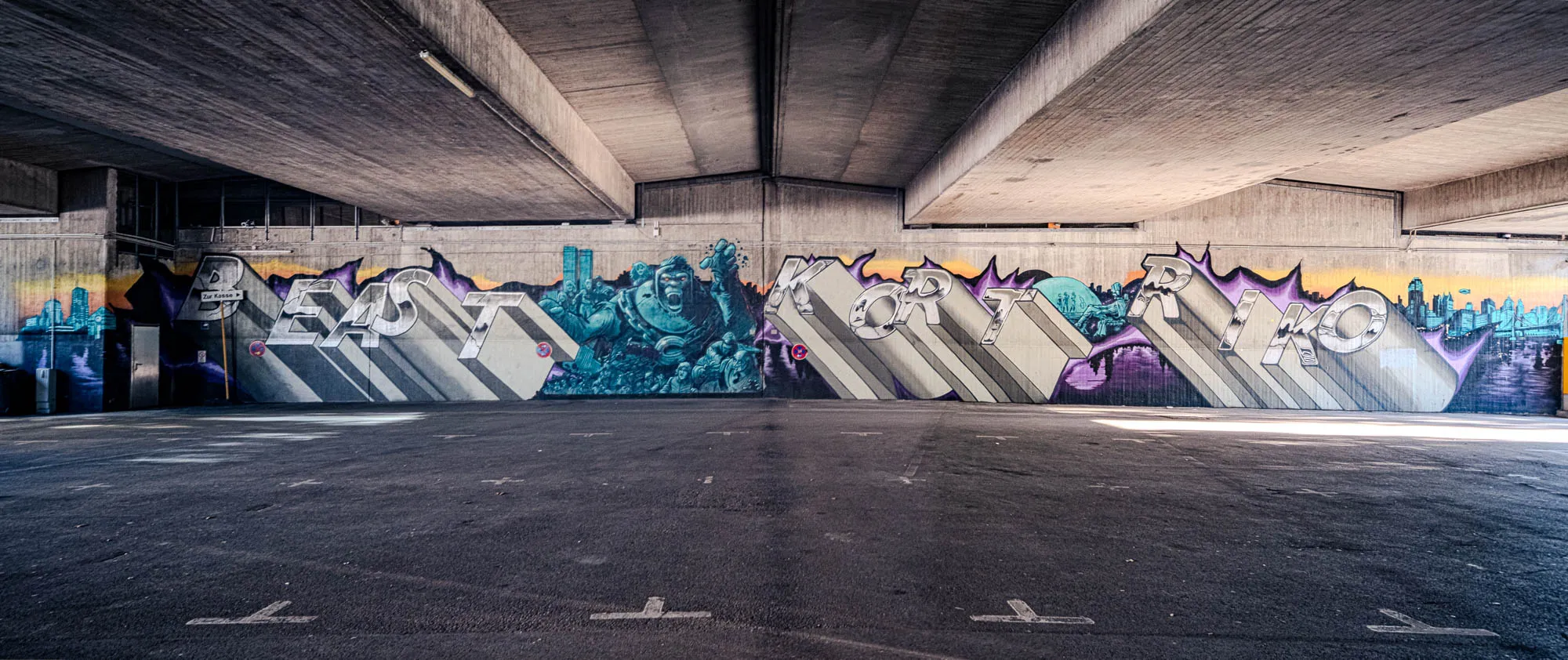The image shows a wide-angle shot of a large graffiti mural under what appears to be a concrete overpass or bridge. The mural spans the length of the wall, filling most of the view. The foreground is a dark asphalt surface, likely a parking area or road, with subtle markings on the ground. The massive concrete structure above is composed of several thick, horizontal slabs that converge towards the center of the frame, creating a sense of depth and weight. The graffiti itself is vibrant and detailed, featuring three distinct sections. On the left, bold, three-dimensional lettering reads 'BEAST', set against a backdrop of a stylized city skyline at dusk, with shades of orange, blue, and purple blending together. In the center, the mural depicts a blue-toned, imposing gorilla wearing a diving suit and surrounded by a crowd of people. To the right, more blocky lettering spells out 'KORTRIKO' with similar three-dimensional effects, again with skyline elements in the background. The style is urban and gritty, reflecting street art aesthetics. The overall tone is somewhat dark and industrial, contrasted by the vibrant colors and detailed artwork of the graffiti.