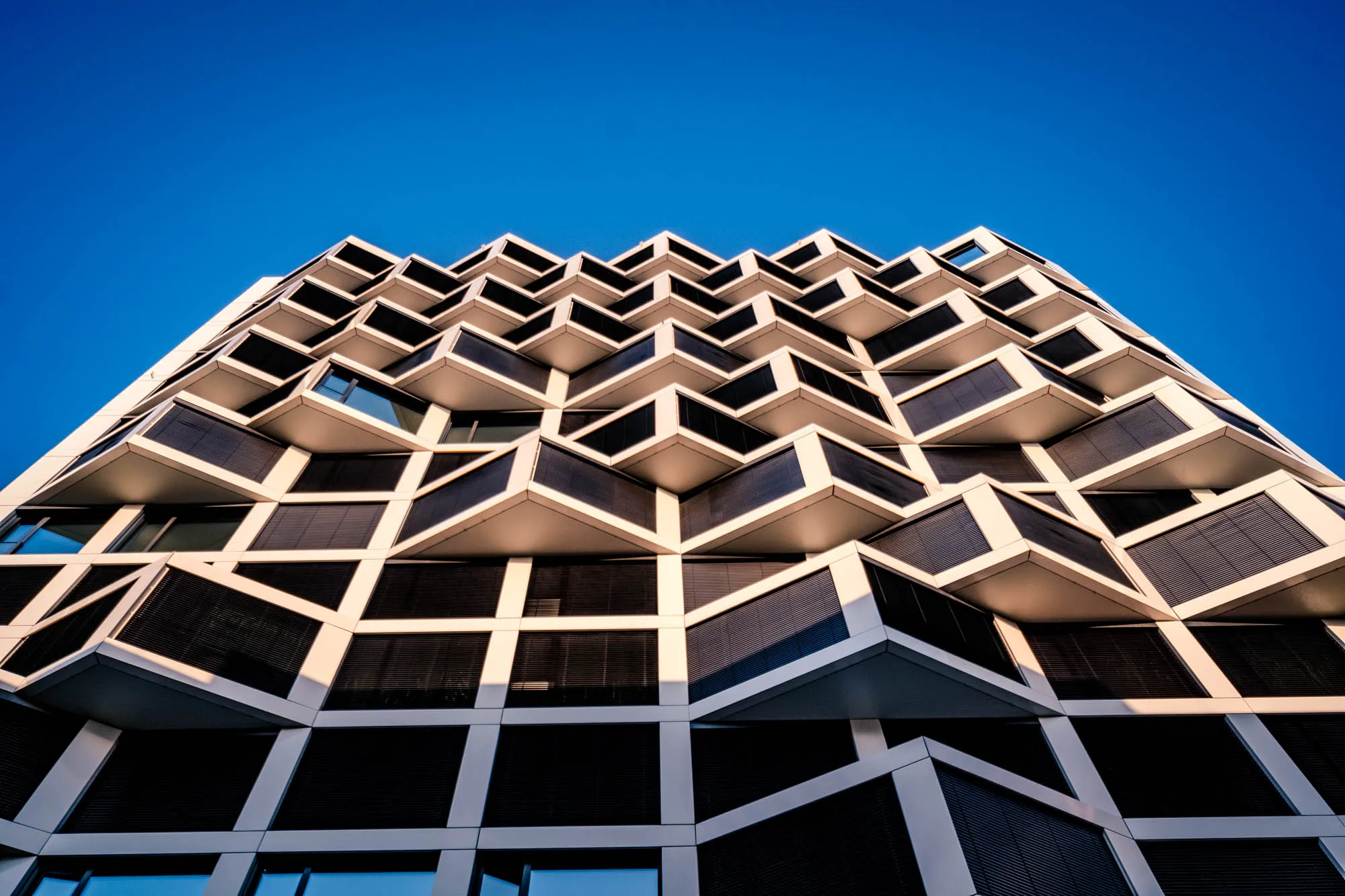 The photograph presents a low-angle, upward perspective of a modern architectural building. The building's facade is distinguished by a repetitive pattern of protruding box-like structures, resembling balconies or oriel windows, each housing windows with dark blinds. These boxes are arranged in a staggered fashion, creating a zigzagging effect as they ascend. The building's framework is constructed from white material, providing a sharp contrast with the dark blinds of the windows. The sky, visible in the background, is a clear and bright blue, offering a striking visual contrast to the building's geometric design and color scheme. The lighting suggests a bright, sunny day, with shadows that accentuate the depth and dimension of the building's unique facade.