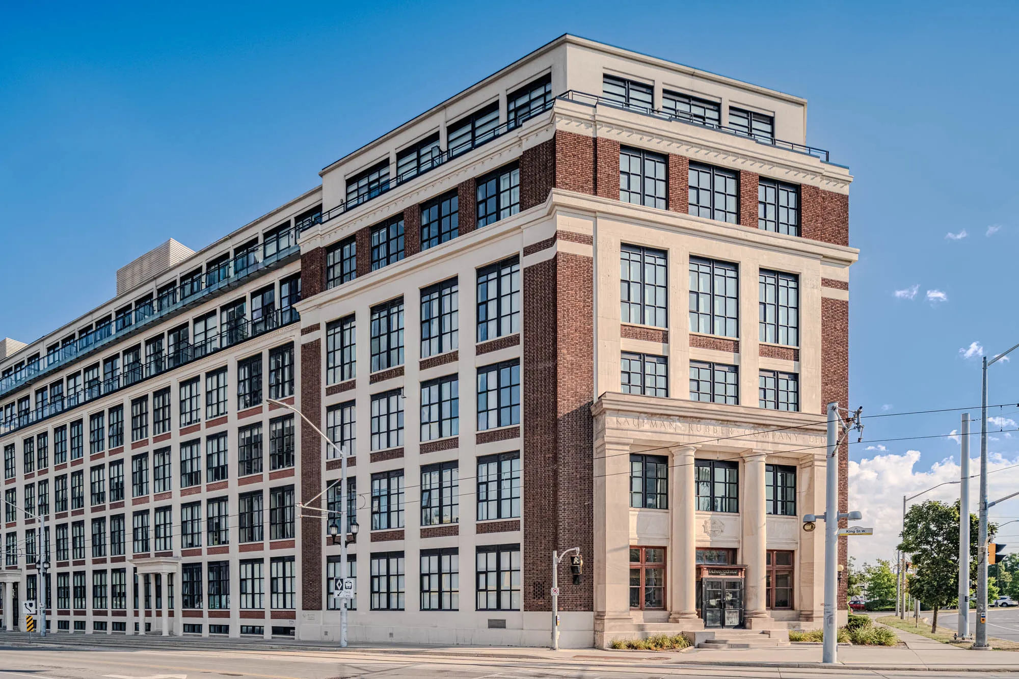 The image presents a stately, multi-story building under a clear blue sky. The building is constructed of a combination of materials: red brick and creamy-white stone or concrete. Large, rectangular windows with black frames line the facade, creating a grid-like pattern. There is an additional wing that appears to have glass balcony fronts for each floor. At the base, a prominent entrance is framed by a neoclassical facade, including columns and a detailed pediment above. The building is situated at a street corner, with a gray sidewalk and street visible in the foreground. Streetlights and signage are present, and a few green trees dot the landscape in the background, to the right of the building, further enhancing the urban scene.