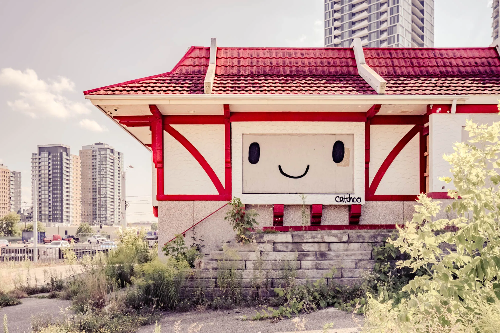 The image shows a small, abandoned building with a whimsical face painted on its facade, set against the backdrop of a modern city skyline. The building is a single-story structure with a distinct Japanese-style roof covered in red, textured tiles. The roof overhangs the building, supported by red beams that create a visually striking geometric pattern against the white, textured walls. The face is painted in simple black lines: two oval eyes and a curved line for a smile. Below the face, the building is elevated on a platform of gray stone blocks, and overgrown weeds and grass obscure the base. The surrounding area appears neglected, with cracked asphalt and wild vegetation. In the background, several tall, modern apartment buildings rise into the pale sky. The overall impression is one of urban decay juxtaposed with playful art, with a soft, muted color palette that lends a slightly faded, vintage feel to the scene.