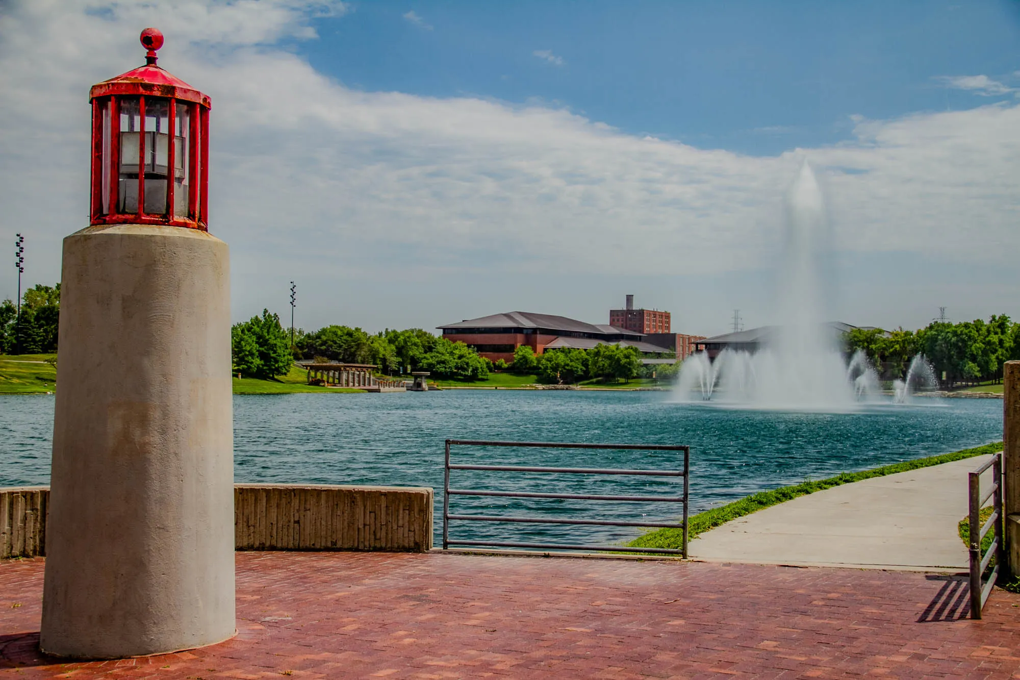 The image shows a tall, cylindrical concrete pillar with a red, metal lighthouse-like structure on top. The lighthouse has a clear, glass-paneled body and a red, pointed roof. The pillar is situated on a brick walkway that extends towards a body of water. There is a metal gate to the left of the pathway, suggesting a barrier of some sort. The water is a deep blue, and there is a large, multi-jet fountain in the center of the water.  There are trees and a few buildings in the background, along with a clear blue sky and some white clouds. The scene appears to be a park or public space with a serene and peaceful atmosphere.