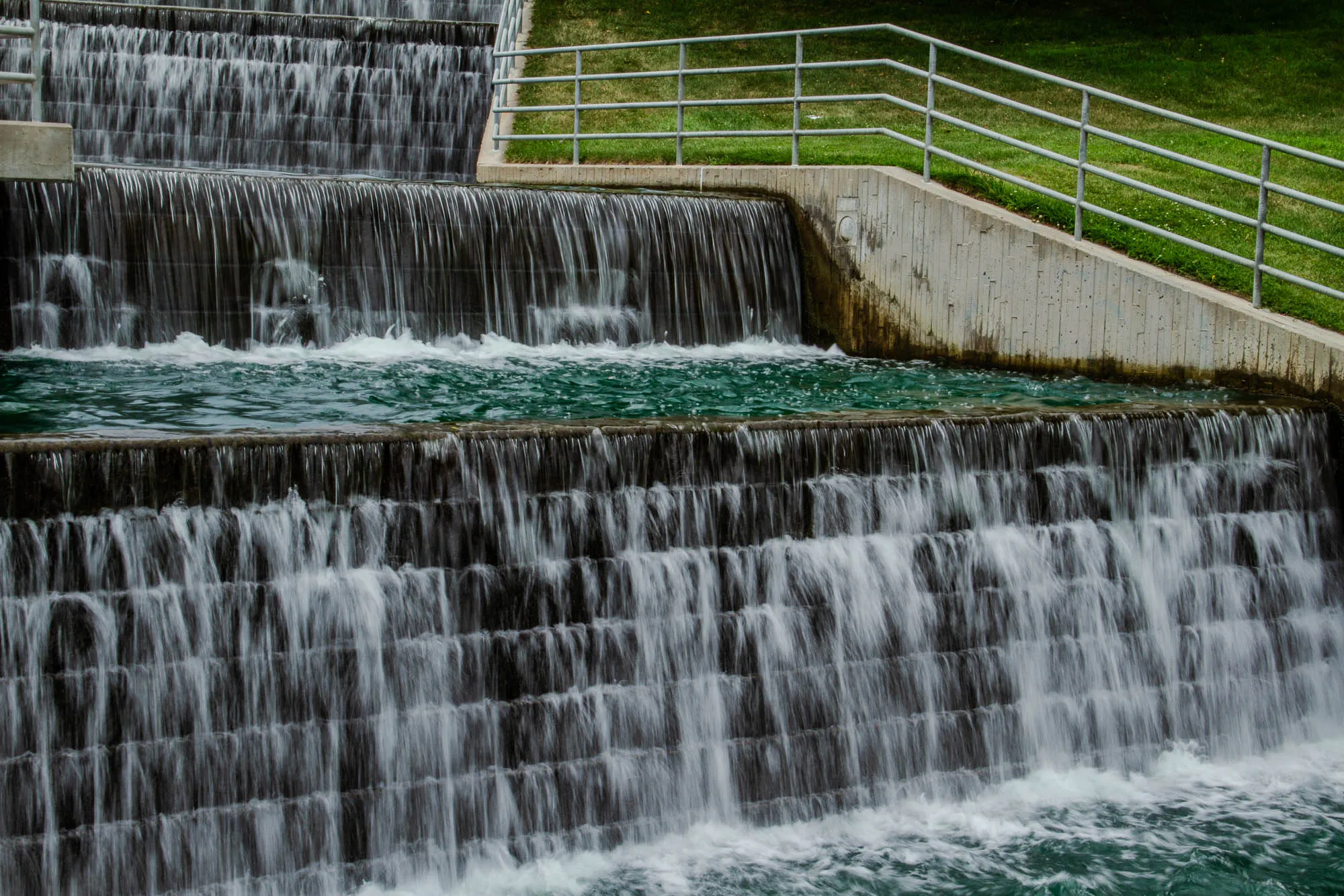 The image is of a waterfall with three tiers.  The top two tiers are made of stone and are quite smooth, you can’t see the individual stones, just the flow of the water. The bottom tier is made of concrete and looks like it has a rougher surface. It is possible to see the individual bricks of this tier.  There is a metal railing and a concrete wall on the right side of the image.  There appears to be a grassy area beyond the wall.  The water is flowing from the top of the waterfall and cascading down to the bottom. The water is clear and you can see the bottom of the waterfall.  It looks like the water is moving quickly. The image is taken from a low angle, looking up at the waterfall. 
