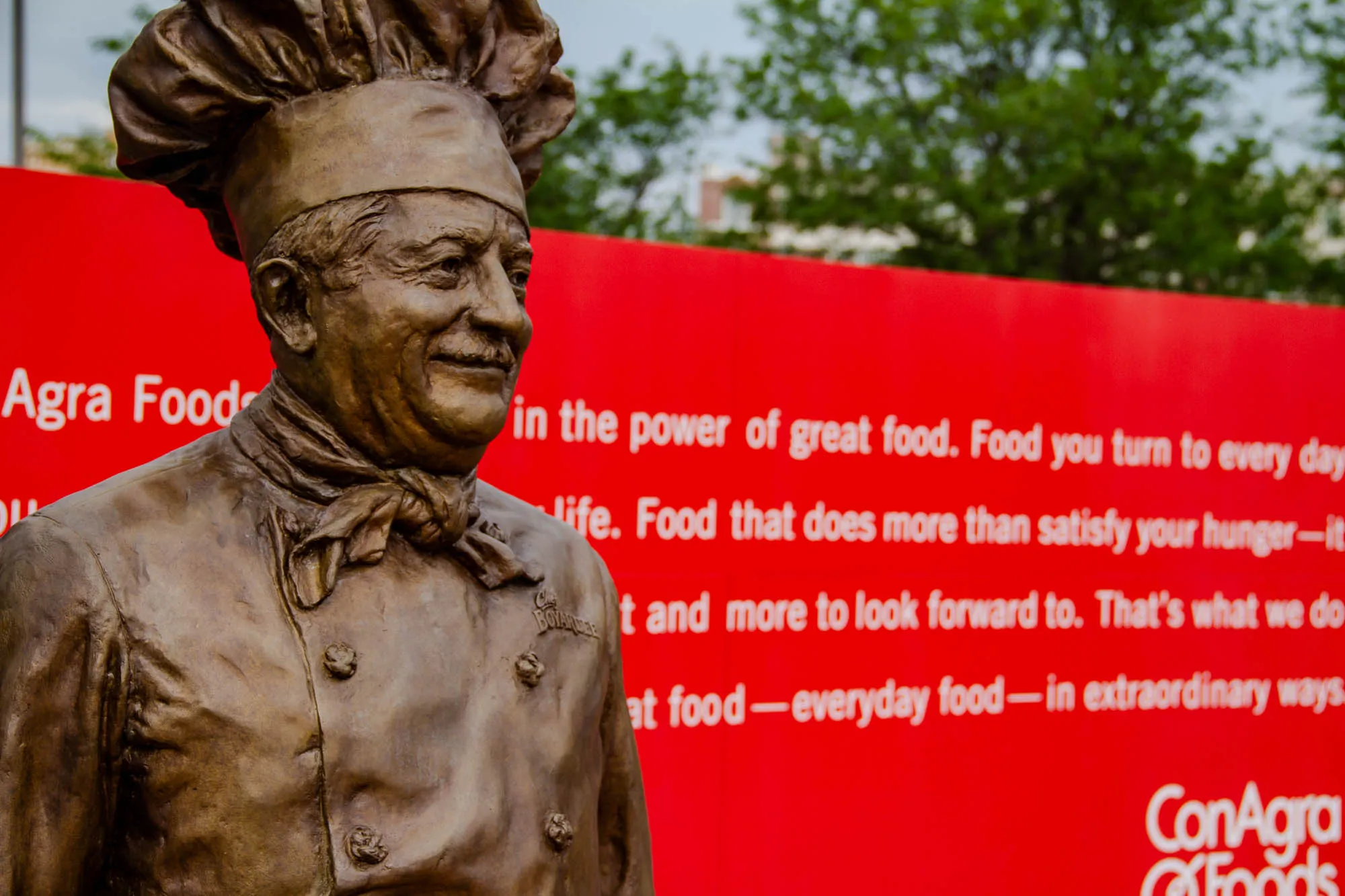 The image shows a bronze statue of a chef, standing in front of a red sign. The chef is wearing a tall, round chef’s hat and a double breasted chef’s jacket. The statue is facing forward, looking slightly to the left, and appears to be wearing a small smile. The chef has a neatly trimmed mustache, and his hair is closely cropped. The statue is a little worn, with subtle details in the jacket and chef’s hat. The red sign behind the statue reads: Agra Food  in the power of great food. Food you turn to every day life. Food that does more than satisfy your hunger-it t and more to look forward to. That’s what we do at food-everyday food-in extraordinary ways. ConAgra Foods. There is a blurred background of leafy trees, with the sky partially visible.  