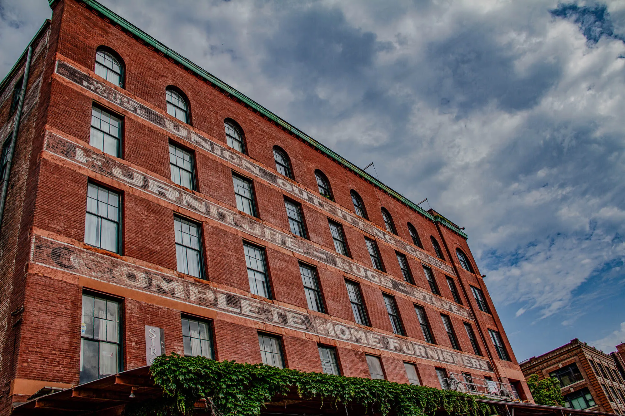 The image shows a red brick building with faded painted advertising on the facade. The building is five stories tall and has large arched windows on the lower stories and smaller rectangular windows on the upper stories. The building is angled upwards towards the sky, which is a bright blue with fluffy white clouds. A few green vines are growing up the side of the building, covering the bottom of the image. The text on the building reads, “Union Outfitters Rugs Stoves Complete Home Furnishers”. The letters are faded and worn, but the words are still legible. The building is clearly old and historic, and it is a reminder of a bygone era. The building is also a testament to the enduring power of advertising.