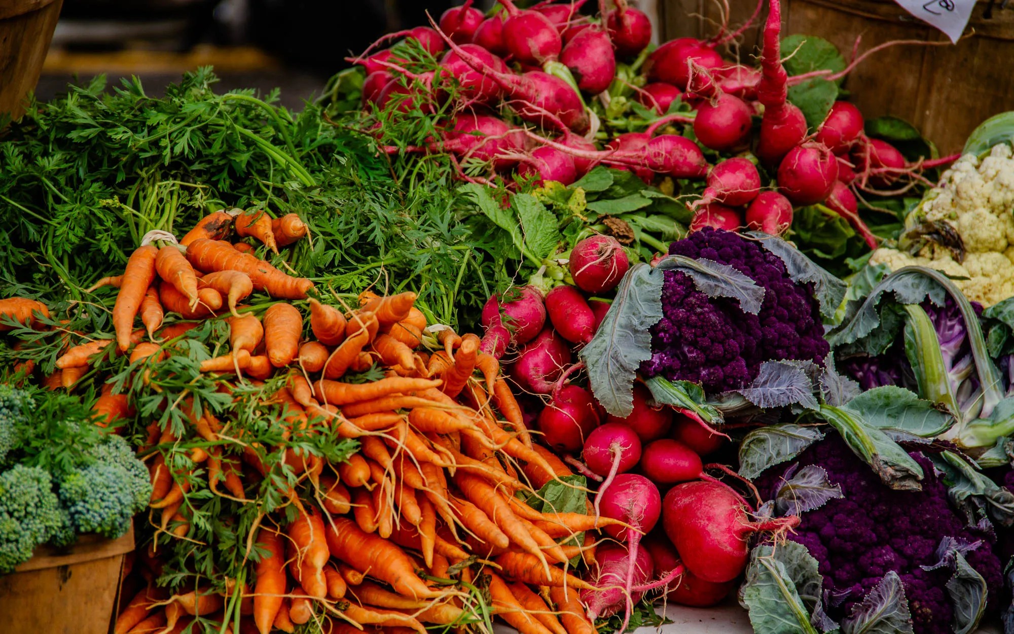 The image depicts a farmers market stall with a variety of fresh produce.  A large pile of orange carrots with their green tops are on the left side of the image.  To the right of the carrots, there are several bright red radishes with green tops.  The radishes are piled on top of a purple cauliflower with dark green leaves.  In the background, behind the radishes, there are some white cauliflower florets. There's also a large bunch of broccoli on the left, in front of the carrots.  The produce is all arranged on a table with a wooden plank in the foreground.  The stall is outdoors, in bright, natural light.