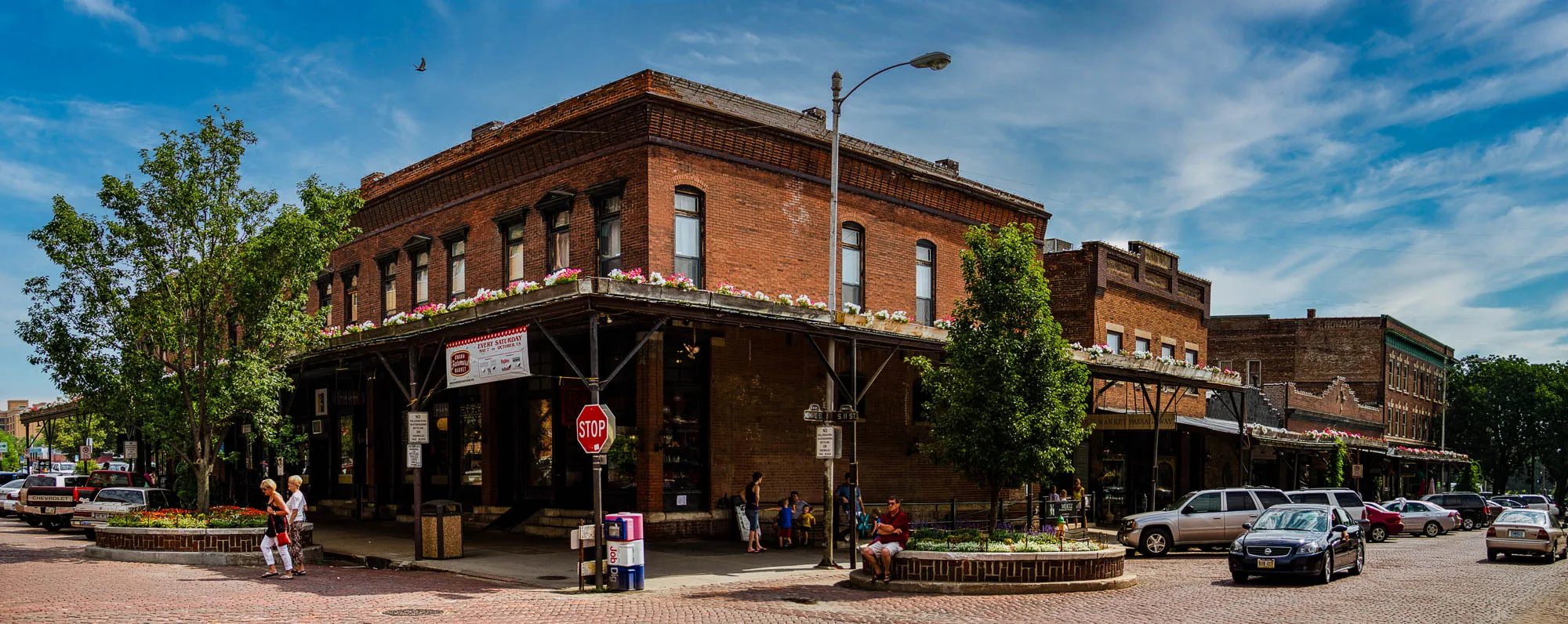 The image shows a brick building with a large awning. It is a market, as a sign indicates "Every Saturday" and "City Market Passaic". A stop sign is visible in front of the building. In front of the building are some flowers and plants. People are walking around the building. There is a street with parked cars and a few people walking on it. The sky is blue with white clouds. There is a large green tree in front of the building and another green tree in the background. There are two women in the foreground walking towards the building. There is a red car parked on the left side of the image. There is a grey car parked on the left side of the image. There is a black car parked in front of the market on the right side of the image. There is a white car parked behind it. There is a white van parked behind the black car. There are a few people walking around the cars and in front of the market. The pavement is brick. There is a streetlamp in the background.