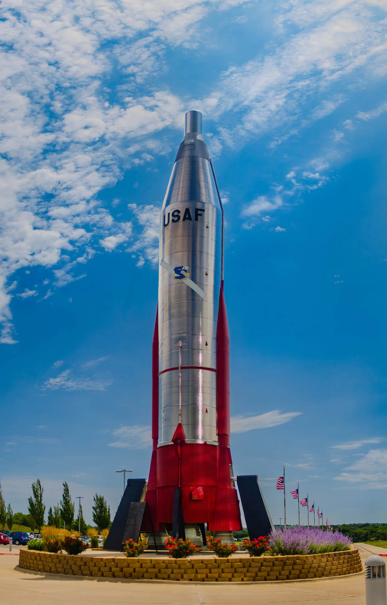 The image shows a large, silver rocket with a red base and a black top. It has the letters USAF written on the side of the rocket in black. The rocket is sitting on a circular brick platform with a low brick wall around it. There is a patch of purple flowers in the foreground, and American flags are lined up in a row behind the rocket. The sky is blue and mostly clear, but there are some white clouds in the background. The rocket appears to be on display in some sort of museum or park.