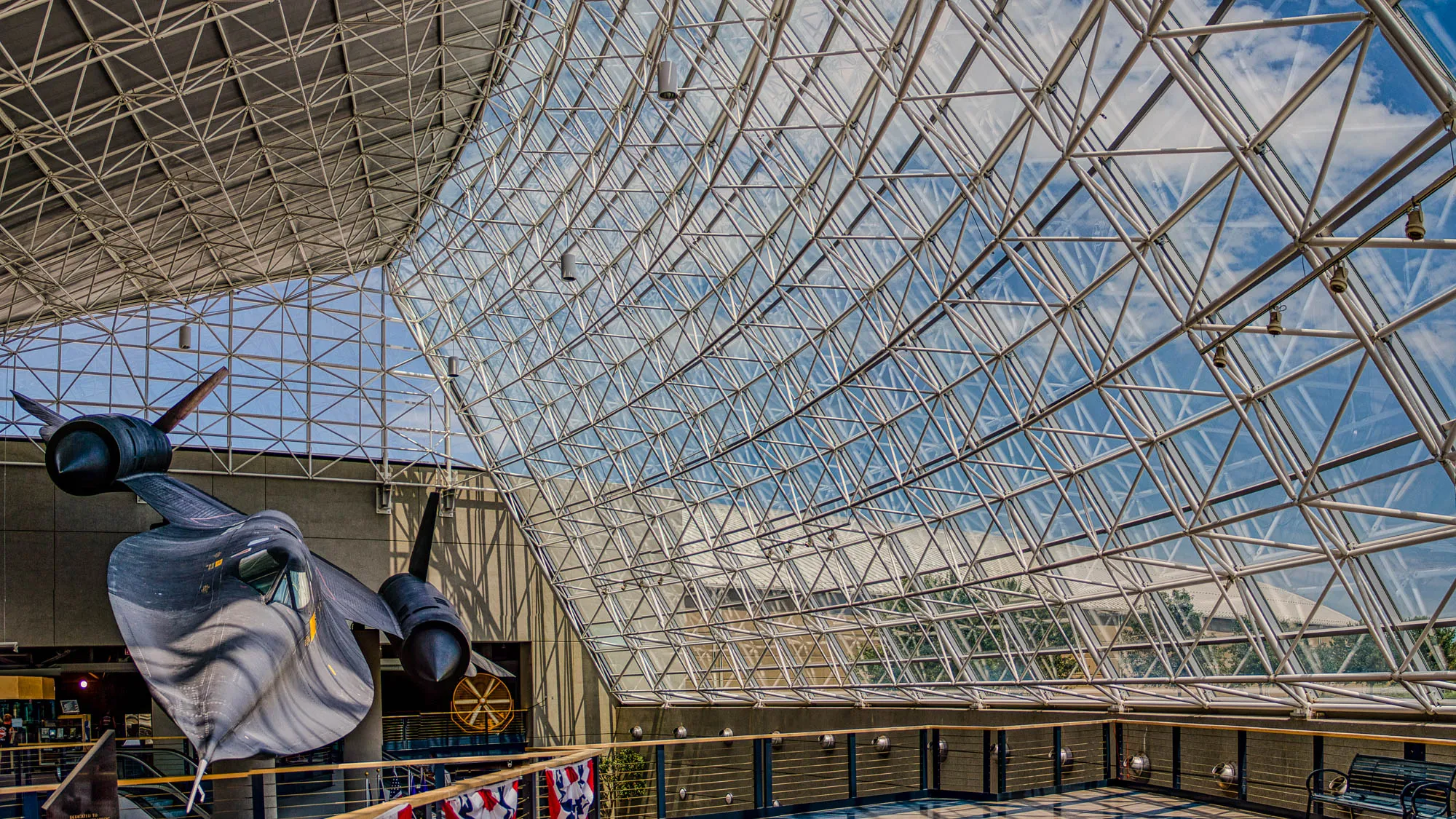 This is a photo of a large black jet suspended from the ceiling inside a building. The ceiling has a large skylight with a geometric pattern of metal beams and glass. You can see blue sky with white clouds through the skylight. The jet is suspended upside down and there are several light fixtures visible in the ceiling.  There is a railing around the edge of the building that looks out onto a sidewalk and there is a metal bench near the railing. The jet is partially covered in shadow. The building seems to be an airport or museum.  The photo is taken from a low angle looking up.