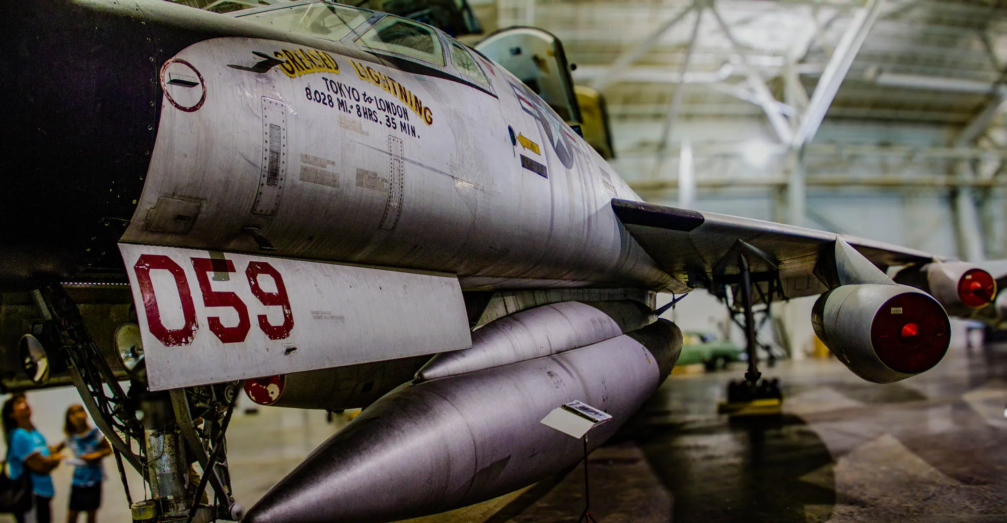 This is a photo of the tail end of a fighter jet. The jet is a gray and silver color, and its tail number is "059." The jet is parked inside of a hangar. The text on the side of the jet reads, "GENSED LIGHTNING TOKYO to LONDON 8.028 MI. 8 HRS. 35 MIN." There are two women in the background of the photo. The first woman on the left is wearing a blue shirt and is looking to the right. The second woman is wearing a blue shirt and is looking down at her feet. The hangar is a bit cluttered with some pieces of equipment laying around in the background.