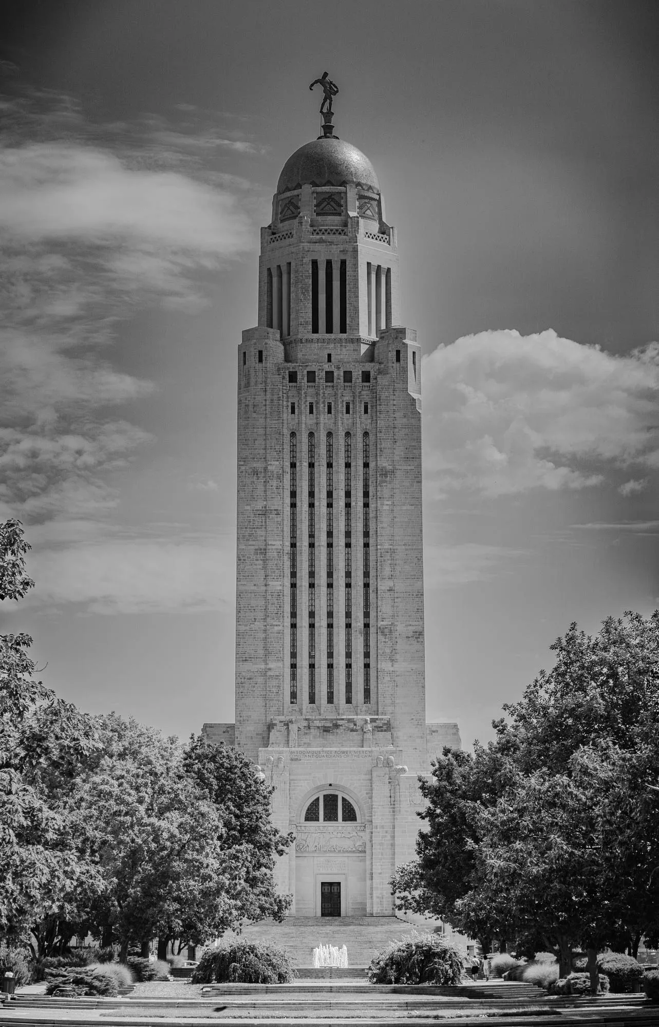 This is a black and white photo of a tall building with a dome on top. The dome is topped with a statue of a man standing with his arms outstretched. The building is tall and rectangular, with many windows running up the side. There are several rows of windows that are grouped together, making them look like vertical strips of glass. There is a stone archway at the base of the building and a fountain in front of it. The fountain has a spray of water coming out of it, creating a mist that appears blurry in the photo. There are trees on both sides of the building, and the sky above is partly cloudy. The building appears to be made of stone, and it has a traditional architectural style. It looks like a government building.