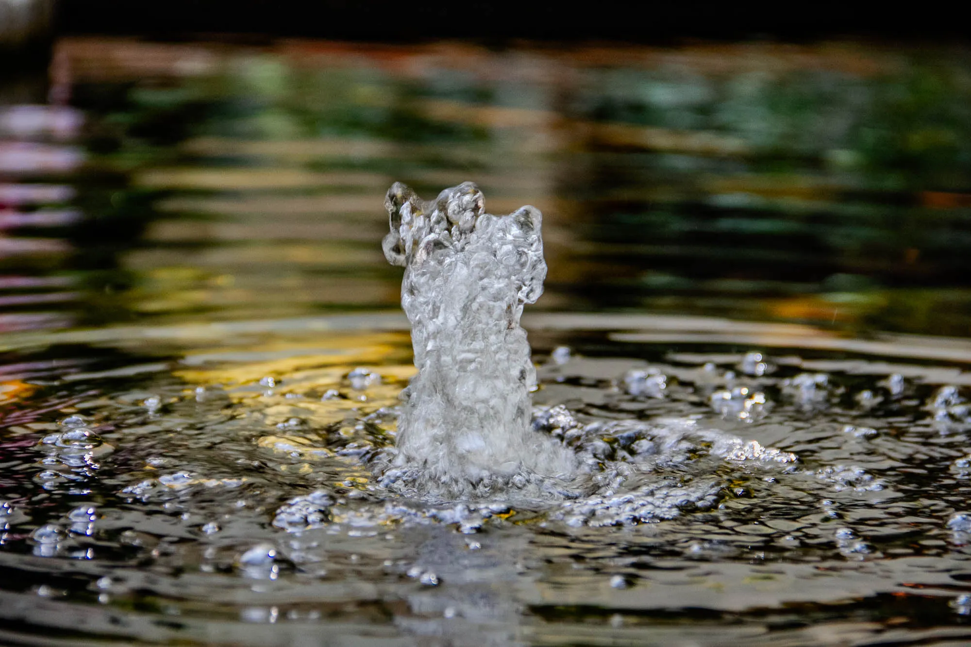 The image is a close-up of a water fountain. In the center of the image, water is bubbling up out of the water, forming a white, billowing column of water. The water is very active and creates a lot of bubbles. The background is blurred and appears to be a dark green, suggesting a pond or other water feature. It is a peaceful image that evokes a sense of tranquility.