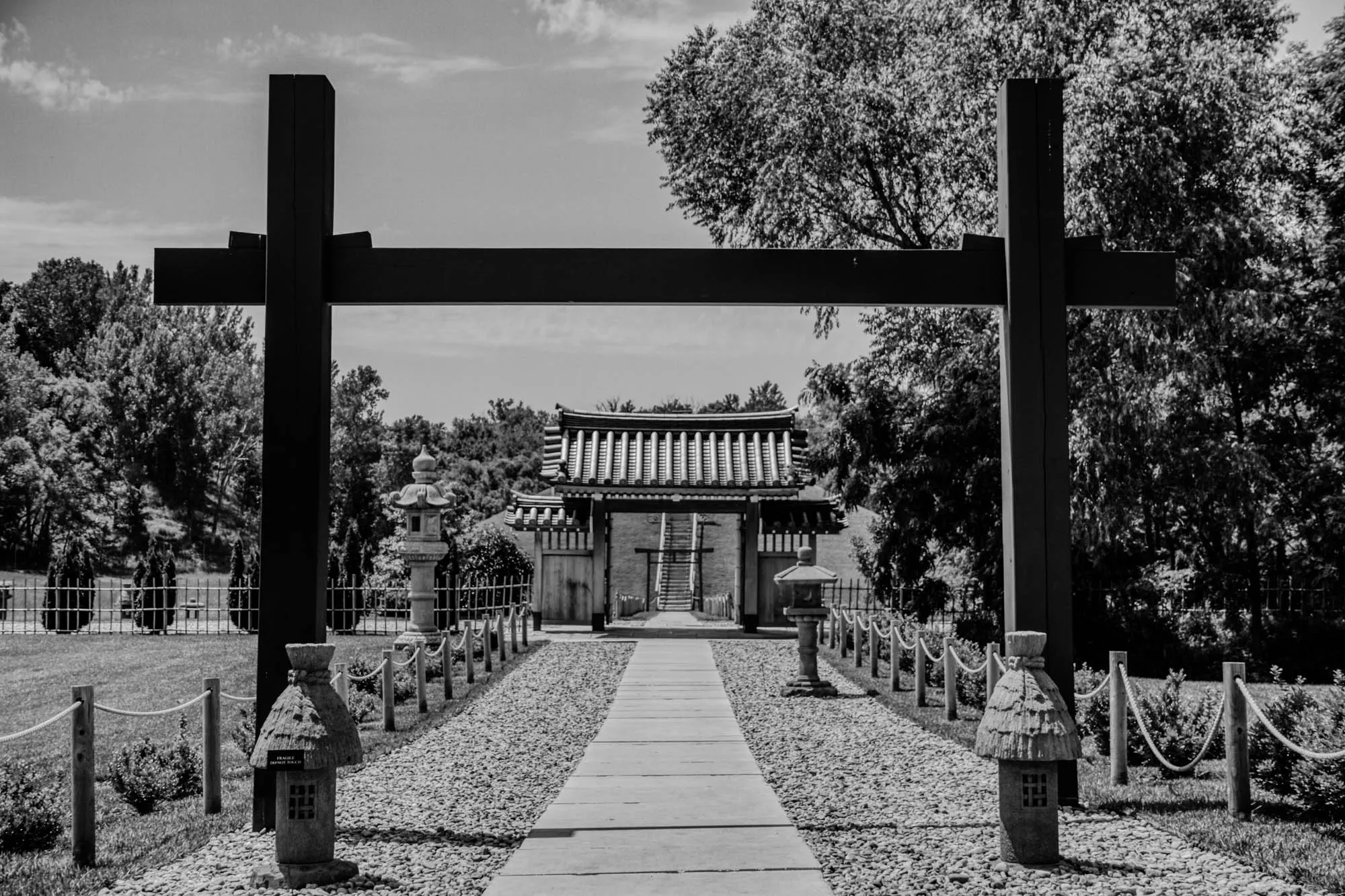 The image is a black and white photograph of a Japanese style garden with a large wooden gate. The gate is made of two tall, vertical posts that are connected by a horizontal beam. The beam has a crossbeam near the top that gives the gate a "T" shape. The gate is framed by trees on either side. There is a path that leads through the gate towards the viewer. The path is lined with small, stone lanterns on either side. The lanterns are about the height of a person's knees and each has a small, rounded top. Behind the gate, there is a small, traditional Japanese building with a tiled roof. It has several doors and windows and is framed by green trees. In the background there is a large patch of lawn bordered by a wooden fence and a row of evergreen trees. The image appears to have been taken on a partly cloudy day. The light in the image is soft and diffused, creating a peaceful and serene atmosphere.