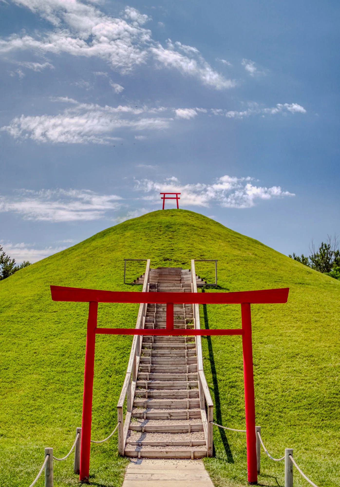 The image shows a grassy hill with a wooden staircase leading to the top.  There are two bright red traditional Japanese gates at the base of the stairs and at the top of the hill. The sky is blue with white clouds. The image evokes a sense of peacefulness and tranquility.