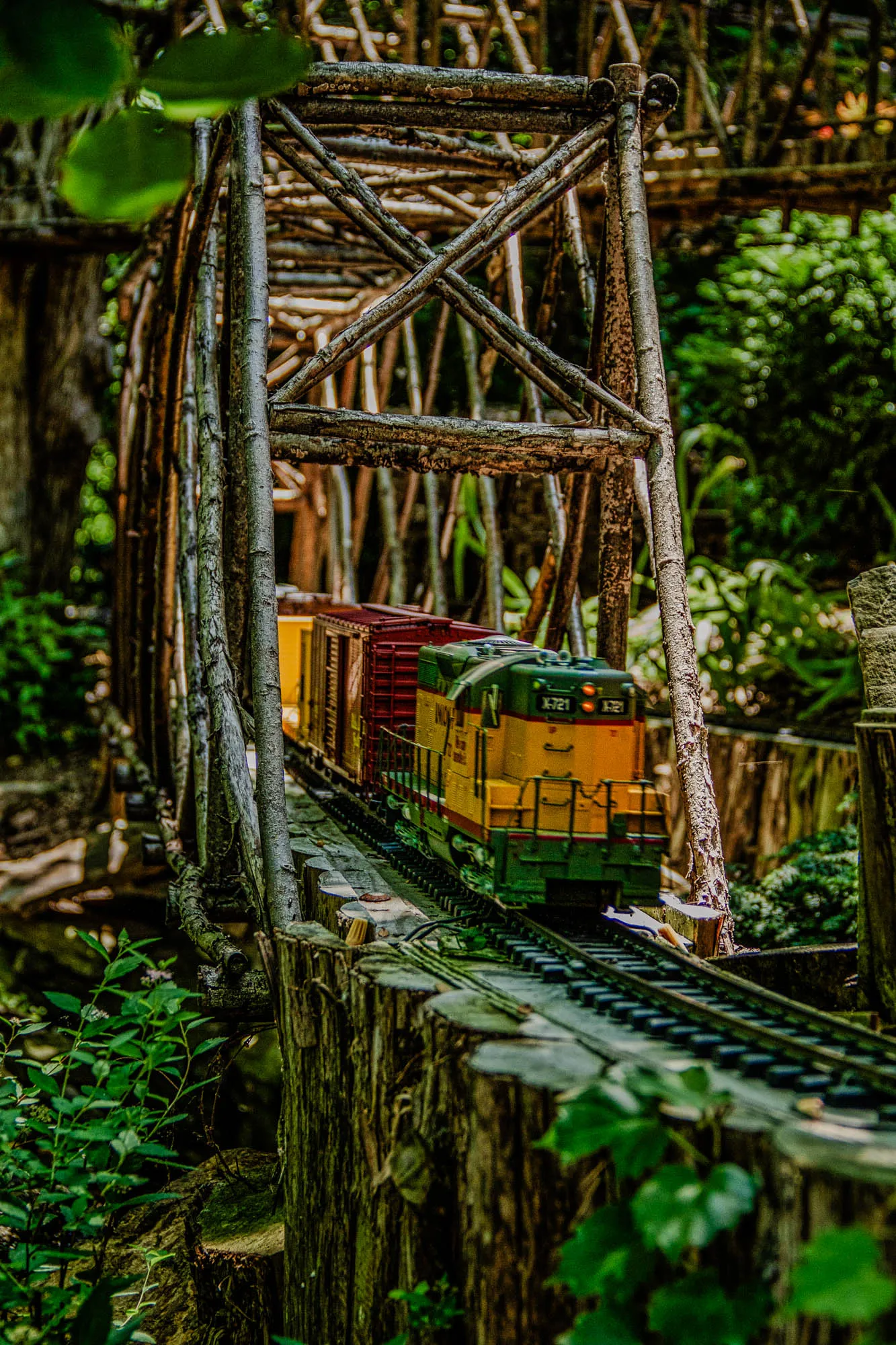 This image is a close-up of a miniature train track with a toy train traveling on it. The train is made of yellow and green plastic and has a red box car attached to it. The track is laid on top of a wooden trestle bridge constructed of large branches woven together in a criss-cross pattern. The trestle is positioned within a natural wooded area with trees and dense green foliage on either side of the track.  The train is in the foreground and moving to the right. The wood trestle bridge is in the mid-ground. In the background, the trees are out of focus.  The image appears to have been taken from a low angle.