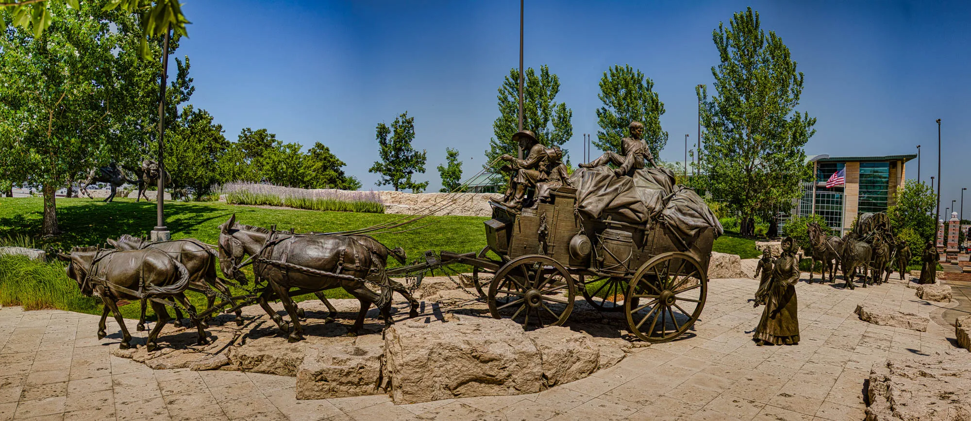 The image shows a bronze sculpture of a wagon train. It's a sunny day, with blue skies and green foliage in the background. The wagon is pulled by six horses, two of which are in the foreground. The wagon is loaded with covered bundles, and a family is riding on the back. Two men are driving the horses. There are several more people walking behind the wagon. In the background, there is a large building with a green roof and a large tree. A small American flag is flying from the building.