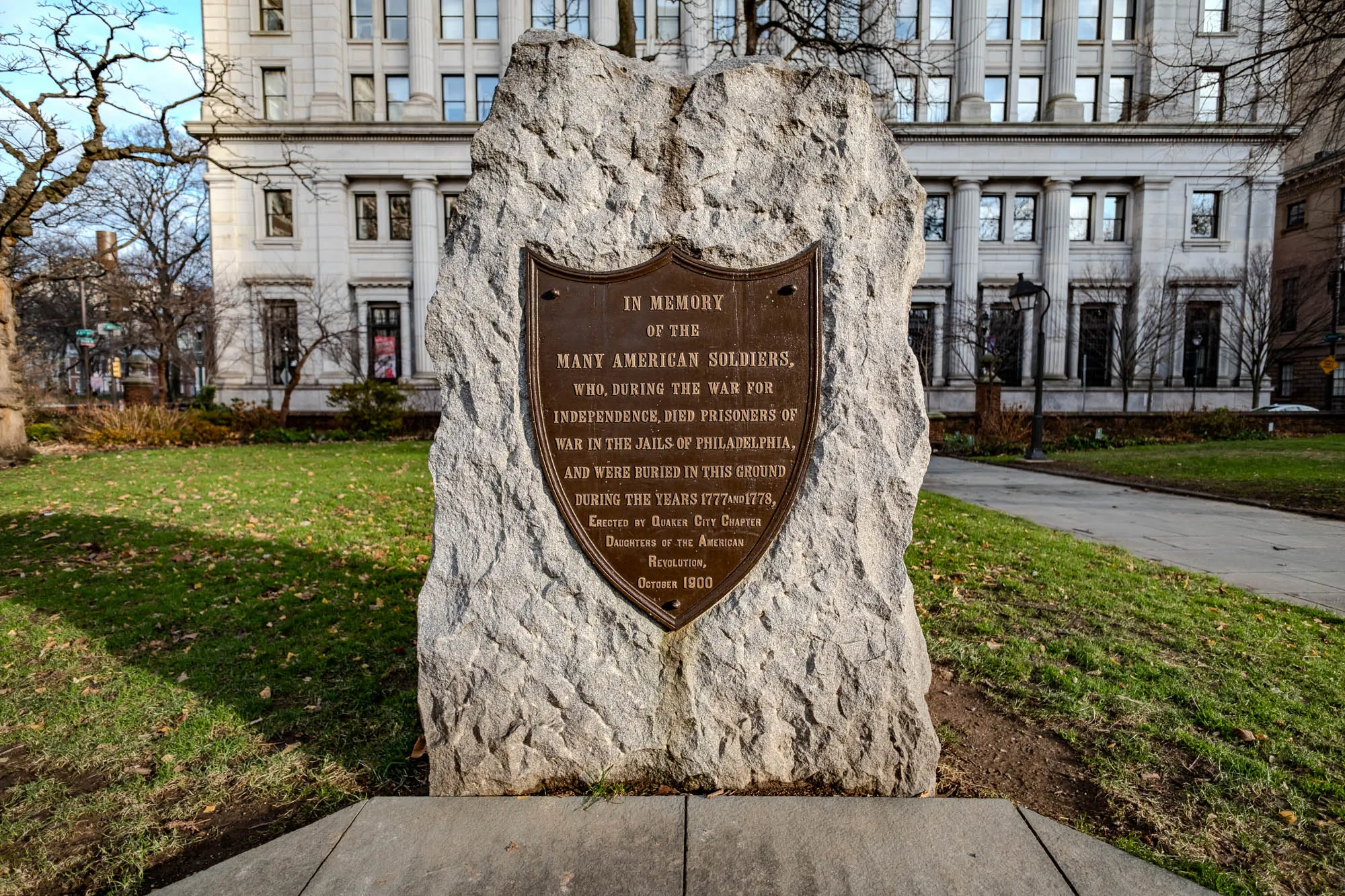 This is a picture of a stone monument, sitting in a park in front of a large building. The monument is a large, gray rock. There is a bronze plaque attached to it in the shape of a shield. The plaque has the following inscription: "IN MEMORY OF THE MANY AMERICAN SOLDIERS, WHO, DURING THE WAR FOR INDEPENDENCE, DIED PRISONERS OF WAR IN THE JAILS OF PHILADELPHIA, AND WERE BURIED IN THIS GROUND DURING THE YEARS 1777 AND 1778, ERECTED BY QUAKER CITY CHAPTER DAUGHTERS OF THE AMERICAN REVOLUTION, OCTOBER 1900." The building behind the monument is tall with white walls and many windows. The ground around the monument is a mix of green grass and brown leaves. There is a walkway to the right of the monument, leading to the building.