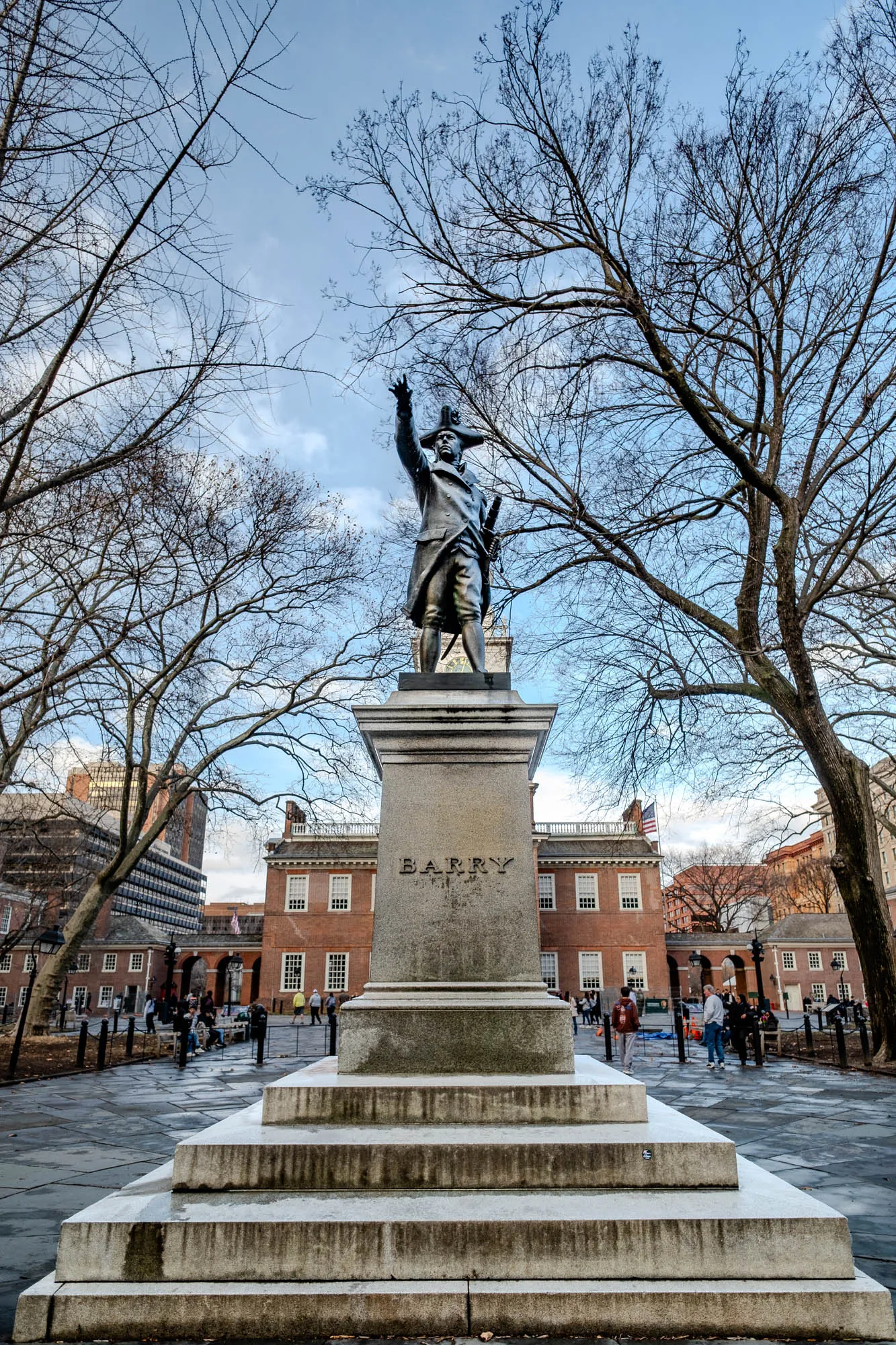 The image shows a bronze statue of a man in a military uniform standing on a stone pedestal. The statue is facing to the right and has its right arm raised in a gesture of greeting or command. It is set in a city park and stands on a square base with the word BARRY inscribed in large letters. There are several steps leading up to the base of the statue. There are two large trees in the background with bare branches. There is a large brick building behind the statue and to the right of the image. People can be seen walking in the distance, and a few people are seated on benches. The sky is a light blue with some clouds.