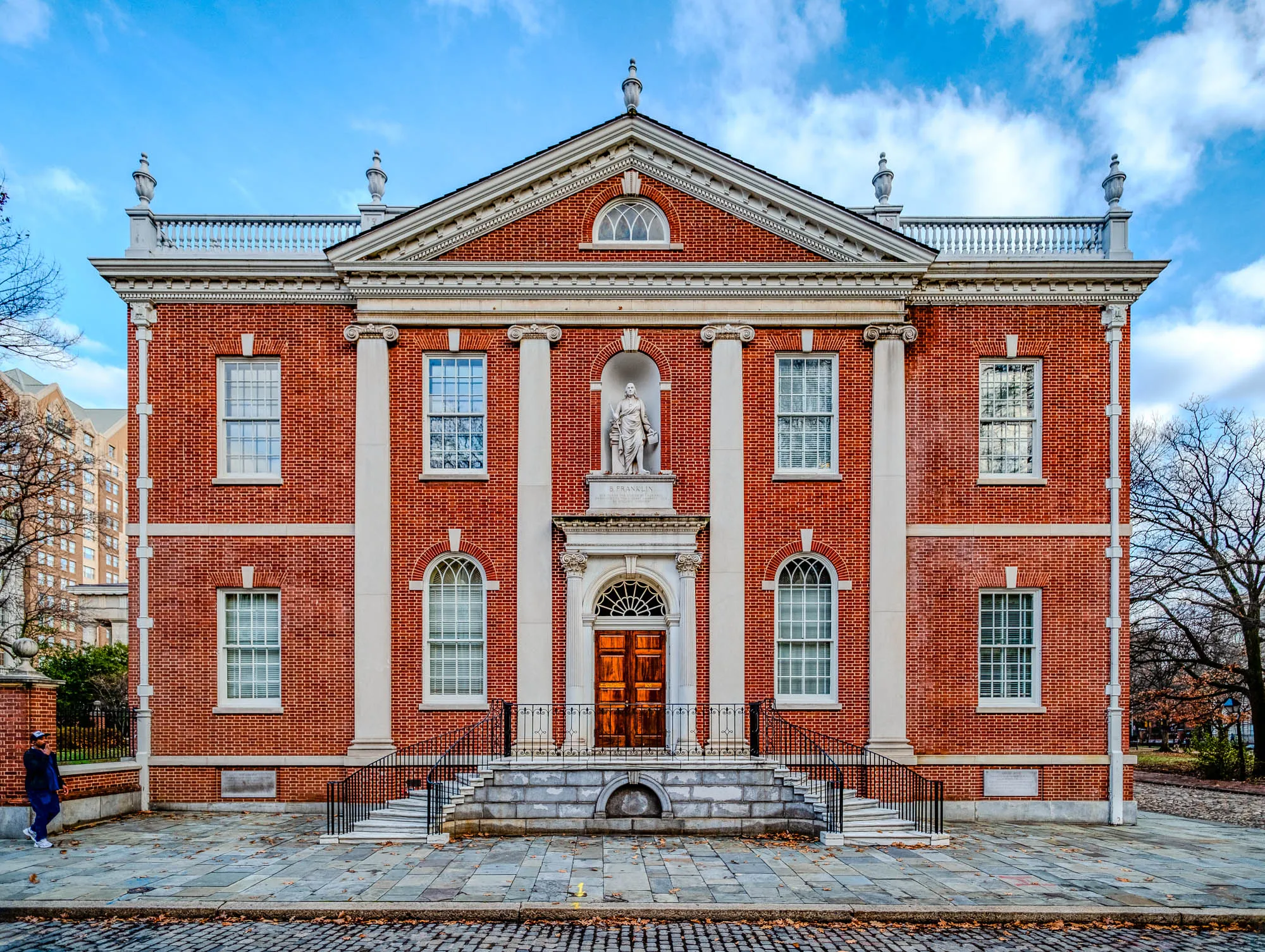 The image shows a rectangular plaque mounted on a brick wall. The plaque is made of dark metal with a thin, light metal border. The inscription reads "AMERICAN PHILOSOPHICAL SOCIETY FOUNDED 1743" in raised lettering. The plaque is centered on the wall, which is made of red brick with light mortar in between the bricks.