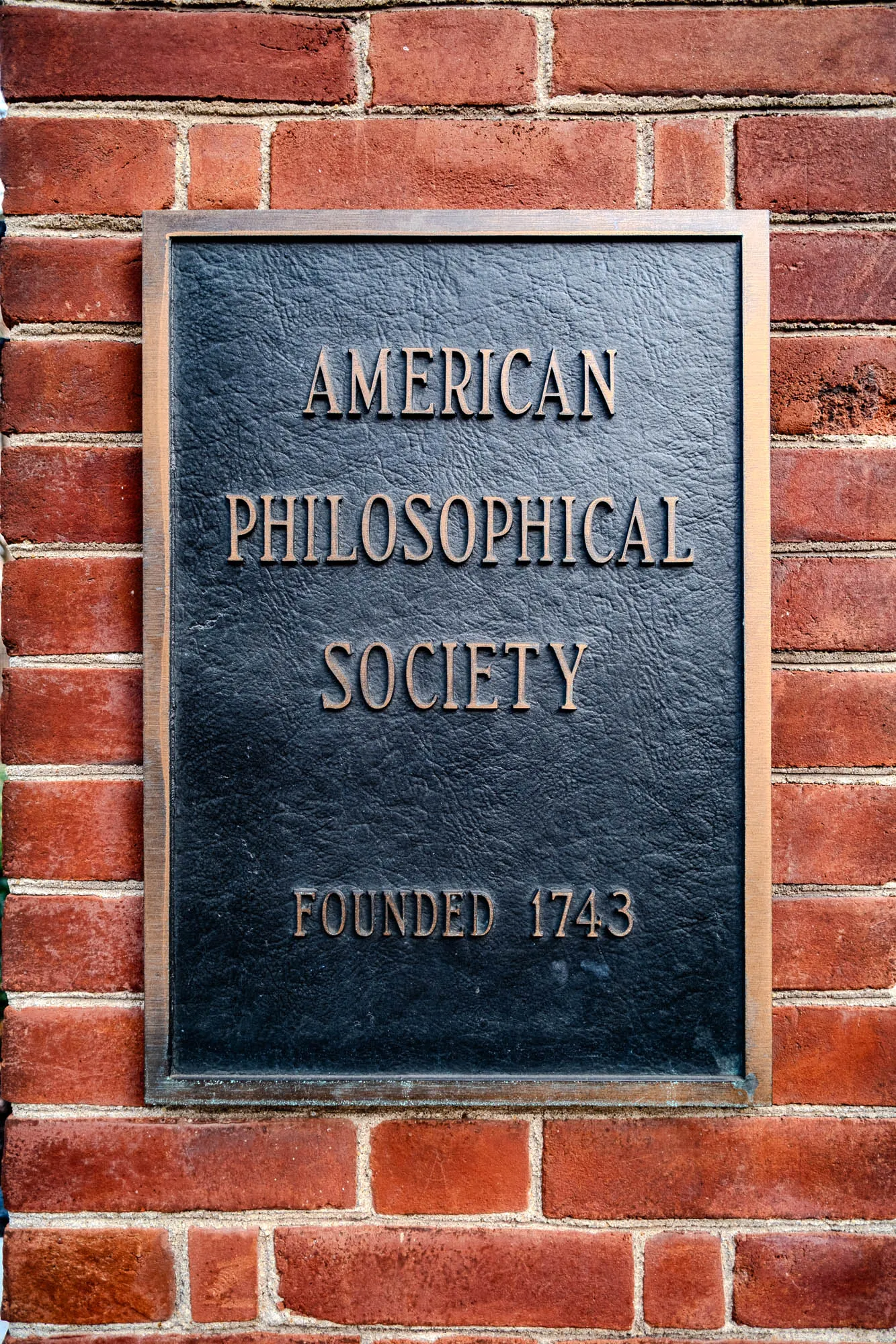 The image shows a black plaque mounted on a red brick wall. The plaque has a bronze border and the following text in raised lettering: American Philosophical Society Founded 1743.  The plaque is rectangular and about two bricks tall.  The bricks are a rusty red with some mortar in between the bricks.  The plaque is slightly to the left of center on the wall.