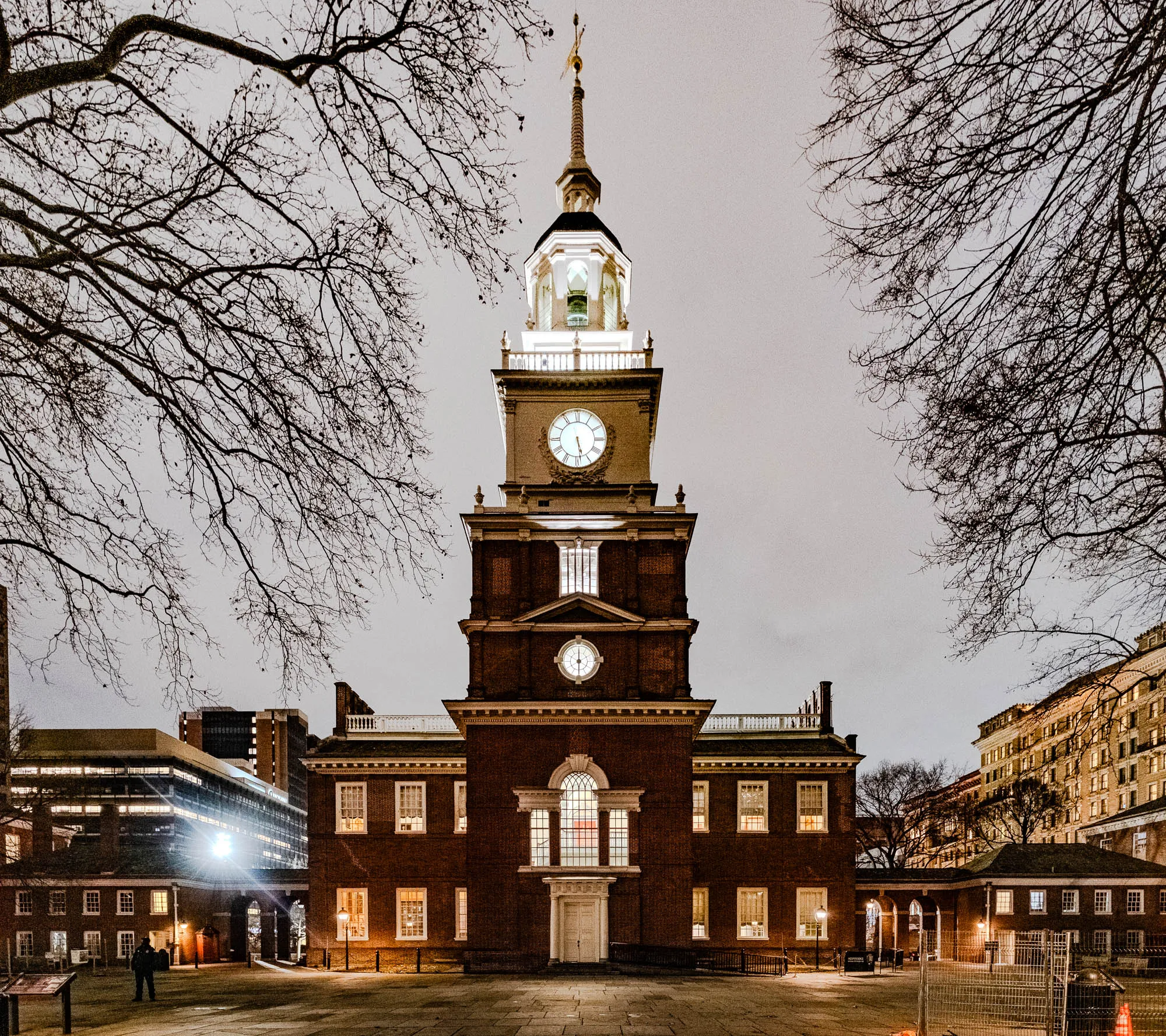 The image shows a brick building with a clock tower, lit up at night. The building is three stories tall and has a tall clock tower with a steeple. The tower has a large clock face on each of its four sides. The building has several windows, many of which are lit up. It appears to be a historic building, maybe a courthouse or town hall. In the background, on the right side of the image, there are more buildings, which are also lit up at night. There are trees in the foreground and background, without leaves, suggesting it is winter time.