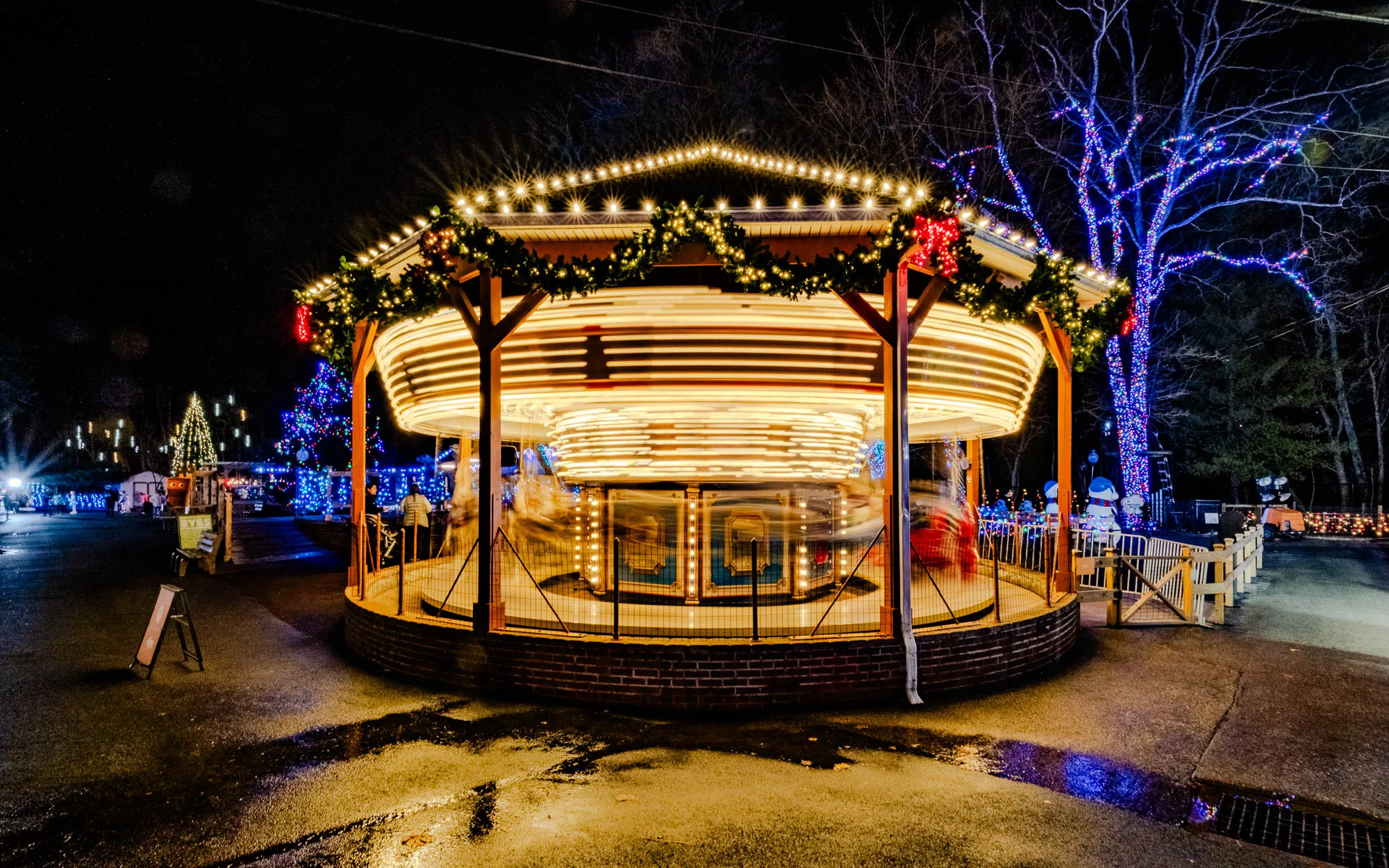 The image shows a carousel at night. The carousel is lit up with white, yellow, and red lights. It is surrounded by a fence and a wooden railing. In the background, a tree with blue lights is visible. The carousel is on a paved surface. The ground is wet and reflecting the lights. The carousel has a wooden roof with garland and lights around it. The carousel is in motion. The lights on it are blurred. There is a Christmas tree behind the carousel, it is covered in blue and white lights. There is a sign on the left that says "EYES".