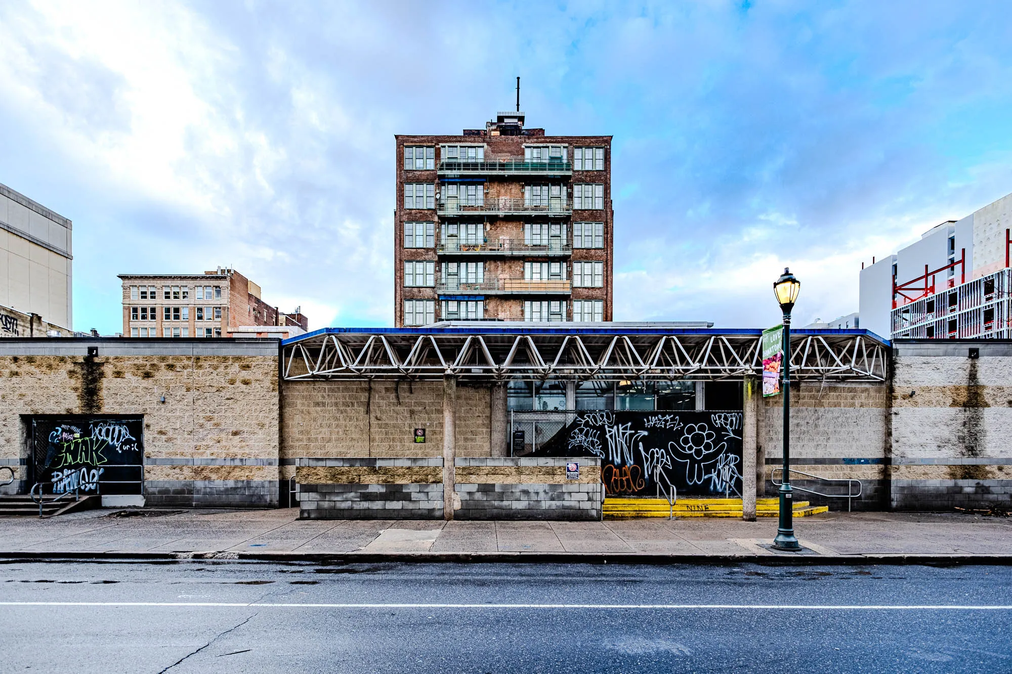 The image shows a street scene with a brick building in the background. The building has many windows and balconies and is a dark reddish brown in color. In front of the building is a brick wall with a metal overhang above it. There are two entrances to the wall, one with a black metal door with graffiti on it and the other with a black metal fence. There is also a street lamp with a round, yellow light on it in front of the wall. The ground is paved and a white line in the road suggests that the scene is likely on a street. The sky in the background is blue with white clouds.