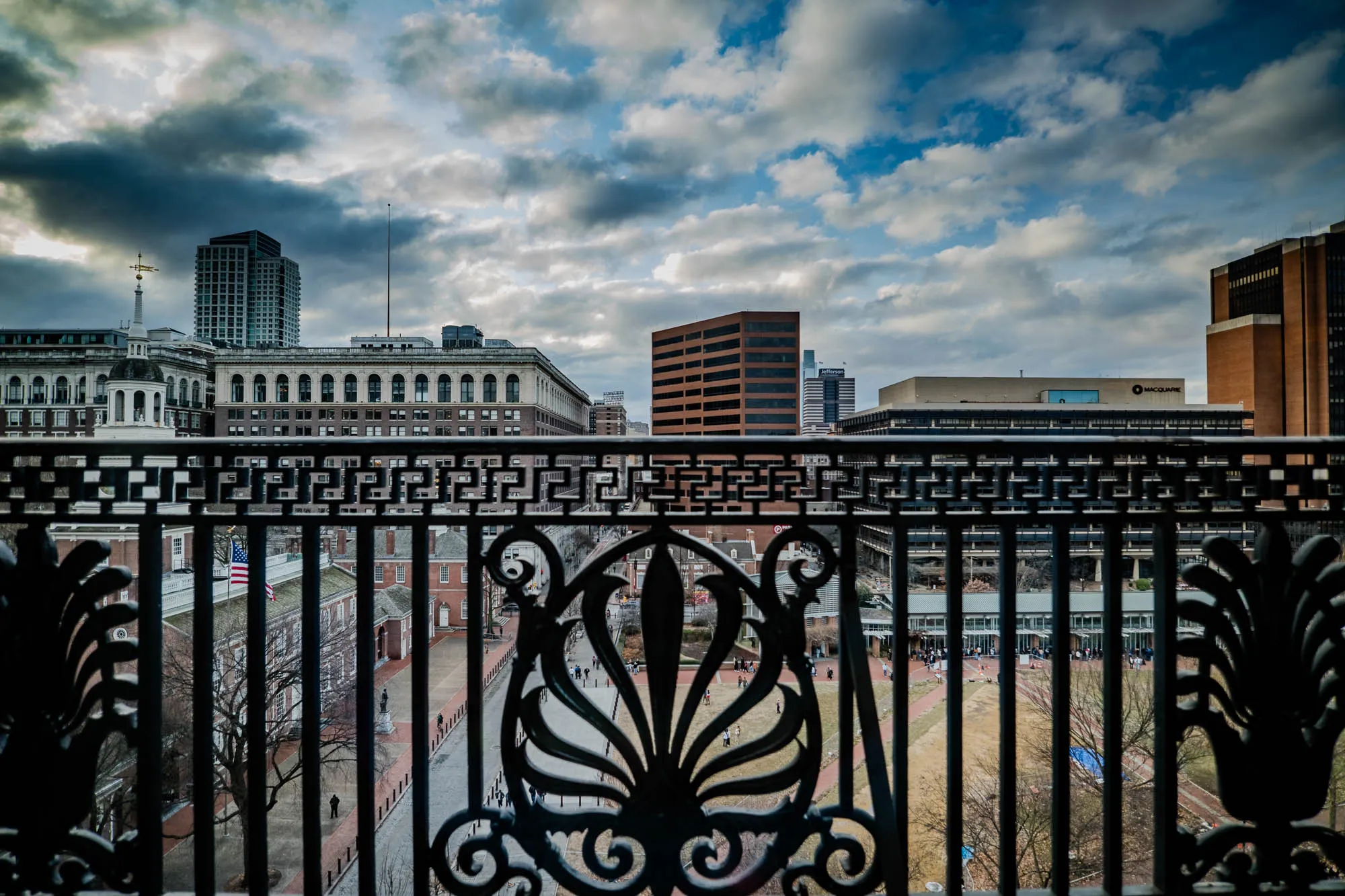 This image shows a view of a city from a balcony, with a black metal railing in the foreground.  The railing has decorative details with swirling patterns and a repeated Greek key design.  Behind the railing is a grassy courtyard with a few people walking around.  In the background is a cityscape with tall buildings, some with modern architecture and some with classic brick facades.  There is a large American flag hanging on a building that is not fully visible.  The sky is mostly cloudy, with some patches of blue. The overall feeling of the image is one of urban life and a sense of history and tradition.