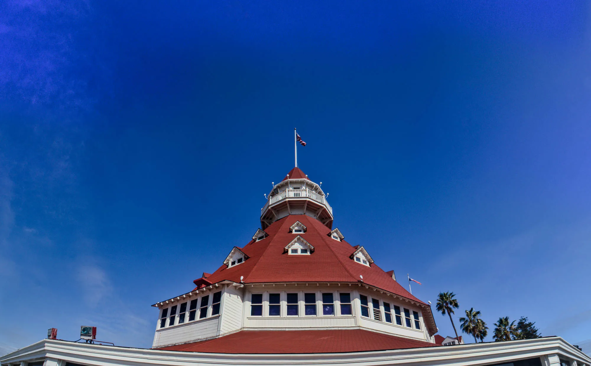 The image shows a building with a red roof against a bright blue sky. There are white trim and windows on the building. The building has a tower at its center with a white railing. An American flag is flying at the top of the tower. The building has a second flag to the right and two palm trees are in front of it. The image is taken from a low angle, looking up at the building. The sky is clear and blue with a few white clouds.