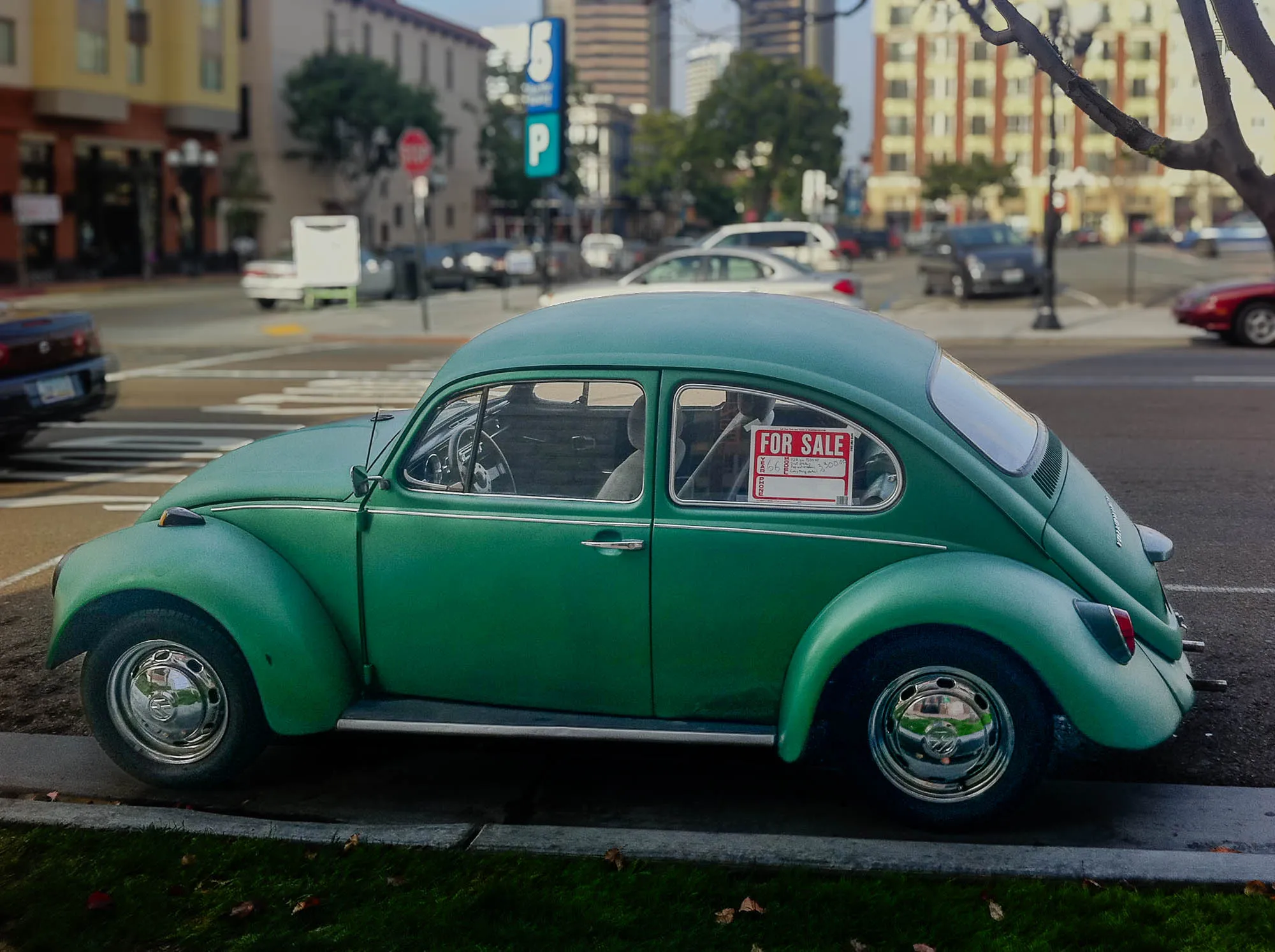 A green Volkswagen Beetle is parked on the side of a street. The car is facing to the right, and the driver's side is visible. The car has chrome hubcaps and a sticker on the back window that reads "FOR SALE". The street is asphalt, and there is a curb and grass in the foreground. In the background, there are buildings and other parked cars.  The photo was taken on a bright day, and the sky is partly cloudy. There is a tree branch in the top right corner of the image.