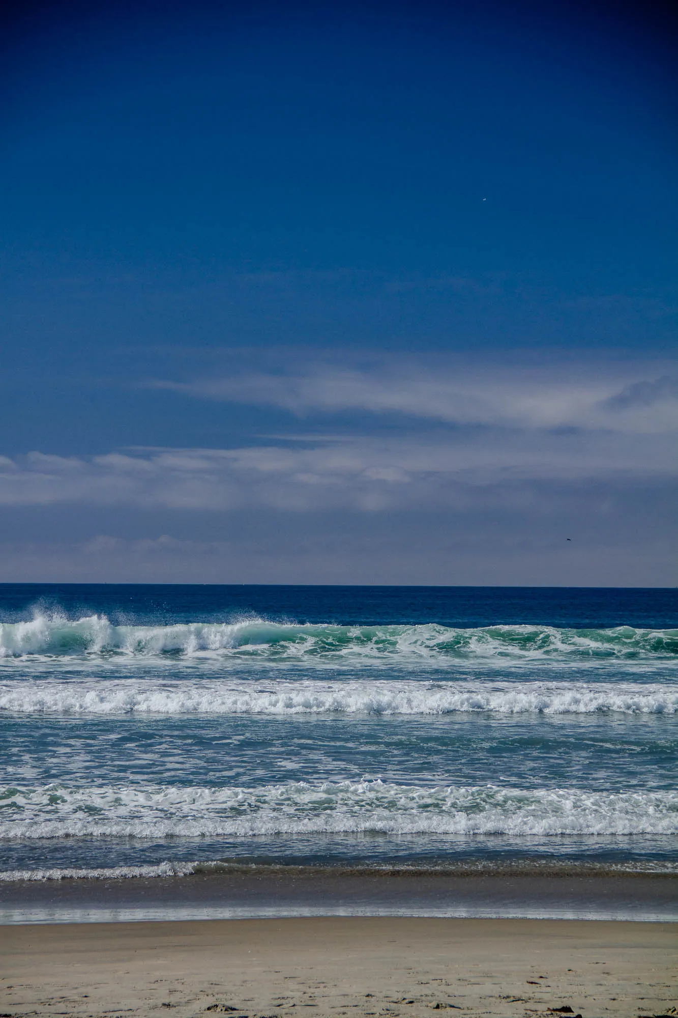 A wide shot of an ocean beach on a sunny day. The light blue sky is mostly clear with some white fluffy clouds. The ocean is a medium blue, with waves crashing on the shore. The sandy beach is light brown and stretches into the distance.  There are some faint footprints in the sand.